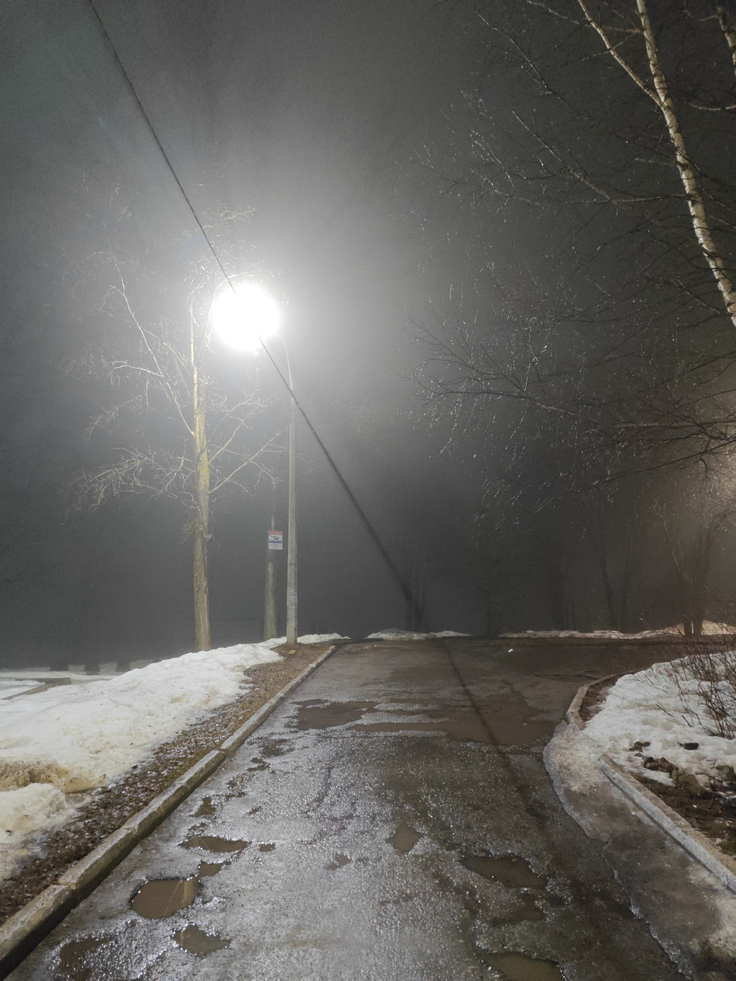The image depicts a street scene in what appears to be the city of Chaikovsky, Russia. It is night and foggy. A street lamp illuminates the scene, casting a bright glow that diffuses through the heavy fog. The road is paved and wet, with numerous potholes filled with water or ice. Patches of snow remain on either side of the road, alongside the curb. Bare trees, typical of winter, are visible in the background, adding to the desolate atmosphere. A utility pole stands near the street lamp. The scene suggests a cold, damp, and possibly late winter or early spring night. There is no evidence of people in the image.