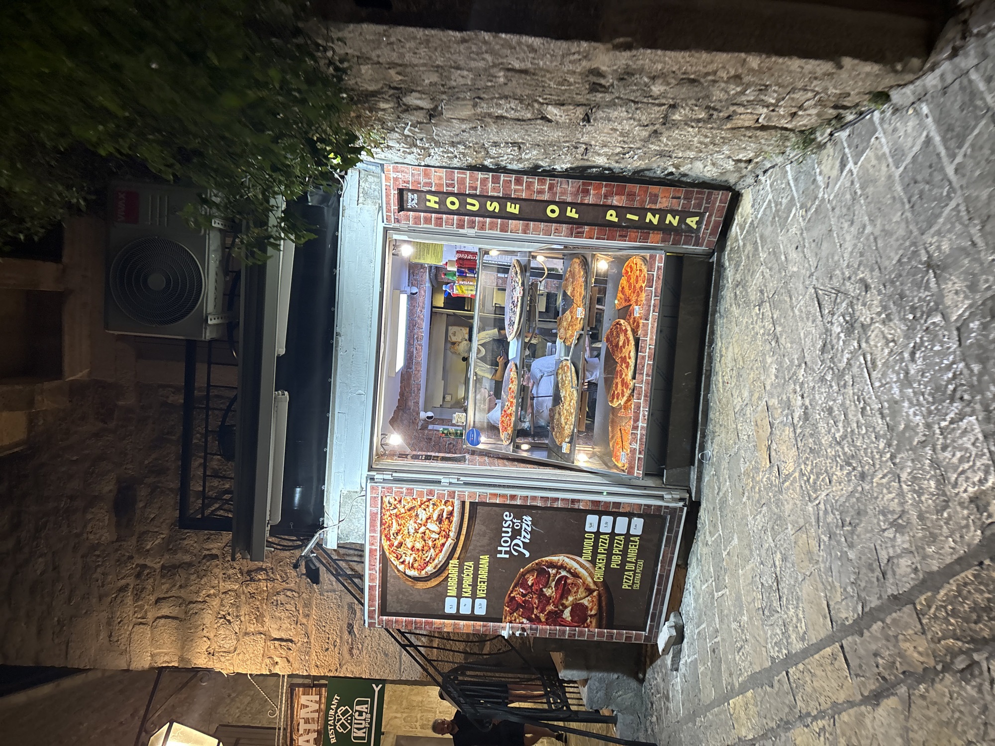 The image shows "House of Pizza," a small pizza shop located on a narrow, cobblestone street in what is likely Budva, Montenegro. The shop is built into the side of a stone building and features a brick facade with the name "HOUSE OF PIZZA" displayed above the serving window. Inside the shop, several pizzas are displayed. A sign on the front of the shop advertises different pizza varieties and prices. A staircase leads up alongside the shop. One person is visible walking up the stairs. The lighting suggests it is nighttime. An air conditioning unit is attached to the wall on the left side of the shop. A sign for a restaurant, "Kuca Pub" is located below.