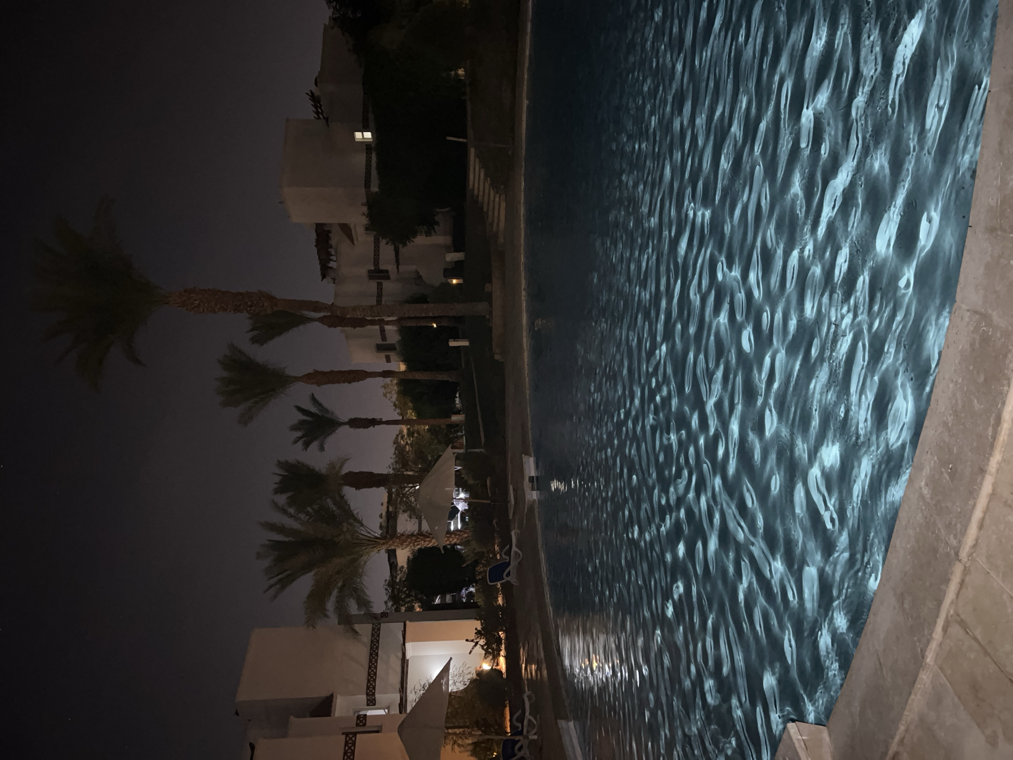 This is a nighttime photograph of an outdoor swimming pool at a resort in Sharm El Sheikh, Egypt. The pool water is illuminated, creating ripples of light on its surface. In the background, there are several buildings with architectural styles common in the region, featuring whitewashed walls and flat roofs. Palm trees are visible, consistent with the desert climate of Sharm El Sheikh. Some lounge chairs and umbrellas are placed around the pool area, suggesting a place for relaxation. The sky is dark, indicating it is nighttime, with a few faint stars visible. The overall scene evokes a tranquil, resort-like atmosphere.