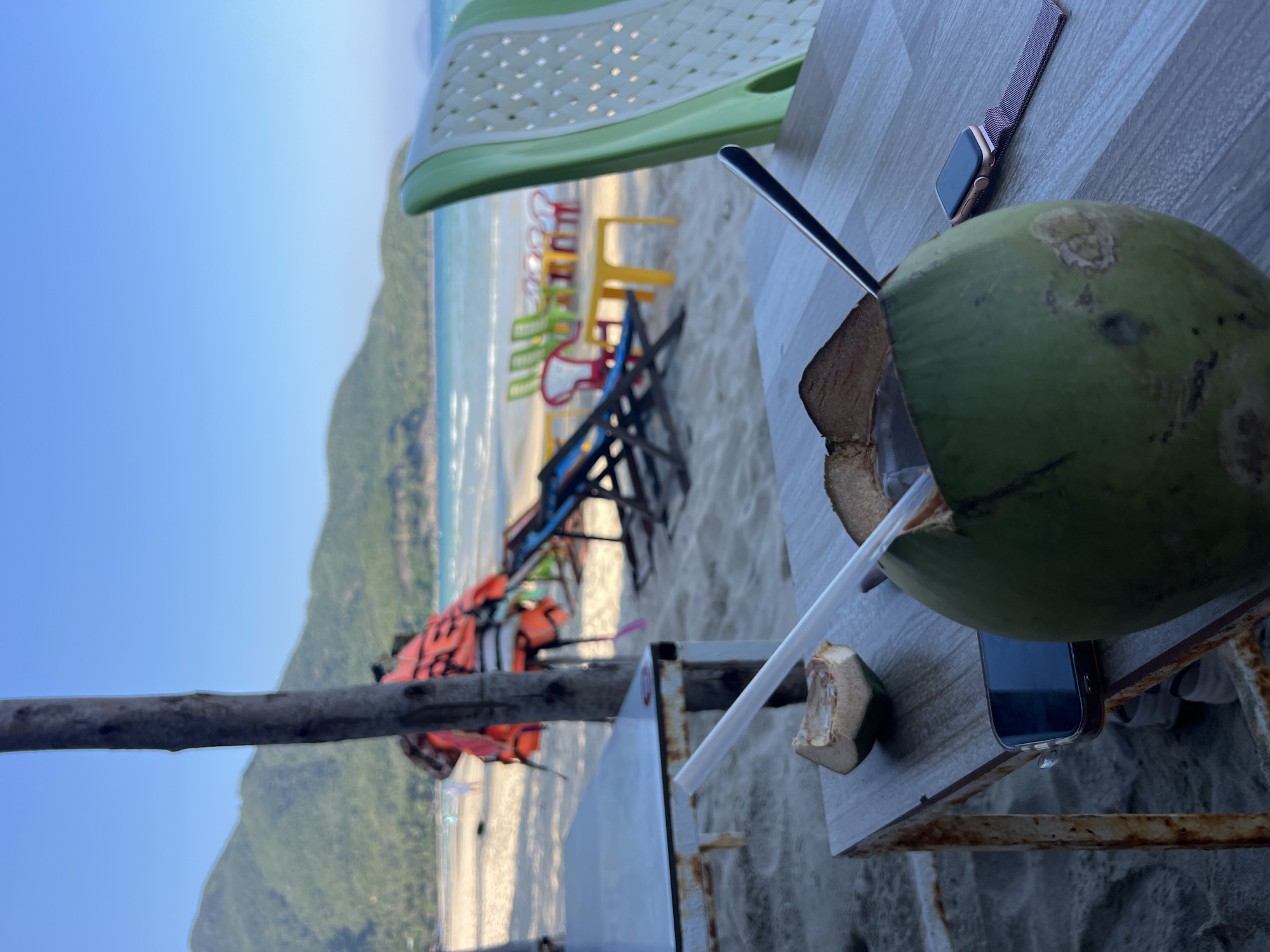 A beach scene in Cam Lâm, Vietnam. In the foreground, a green coconut with a straw and spoon sits on a weathered, light-colored table with a metal frame. An Apple Watch and a smartphone are also on the table. The background reveals a sandy beach with rows of colorful plastic chairs facing the ocean. Beyond the beach, lush green hills rise against a clear, blue sky. Several orange life jackets are draped over an object near the tables.