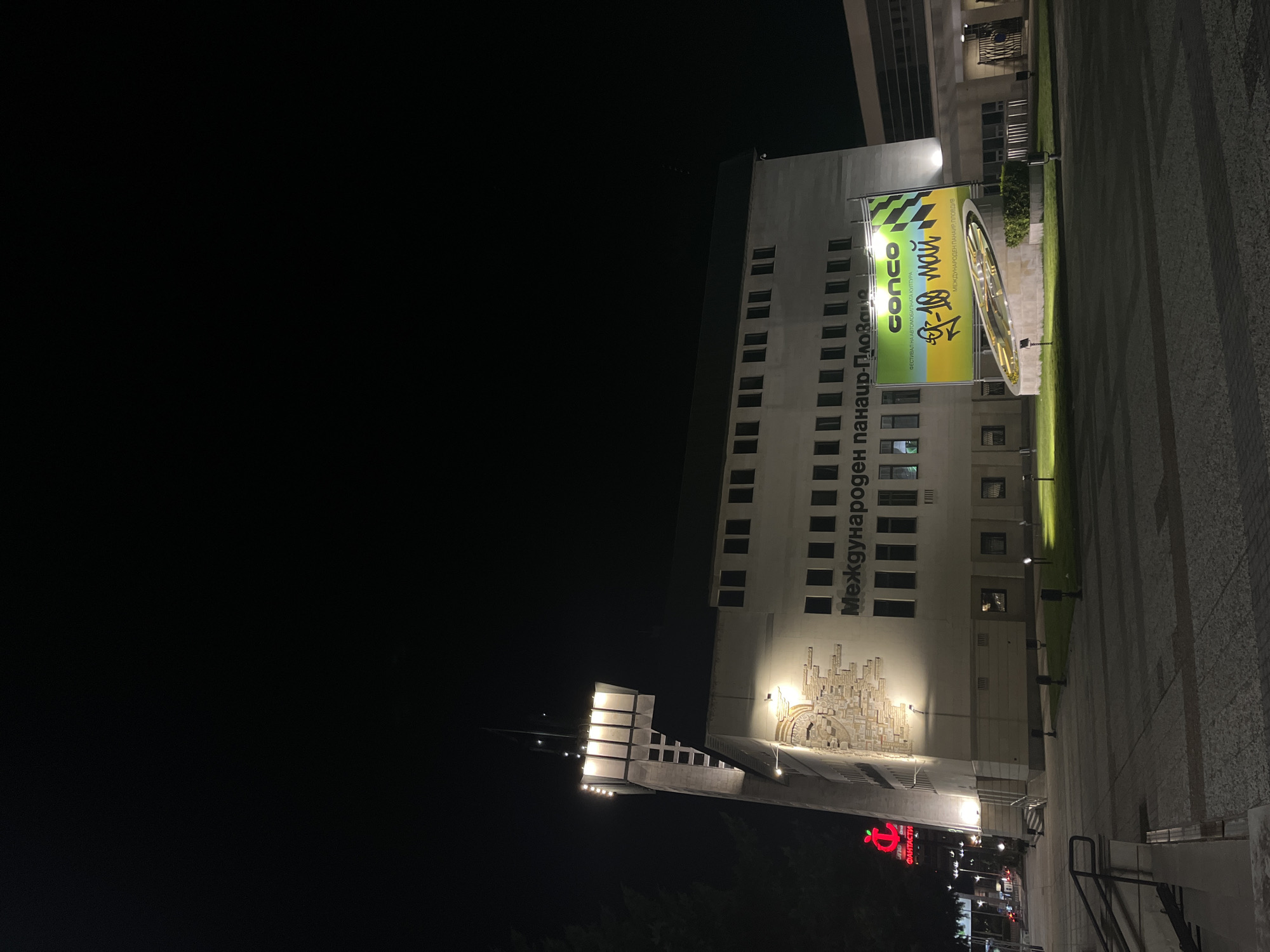 The image shows a view of the Plovdiv International Fair complex in Plovdiv, Bulgaria at night. A large, modern building with multiple windows stands prominently. The building features an architectural ornament under the windows. A billboard advertising "conco", a car culture festival, is next to the building. Streetlights illuminate a paved plaza.