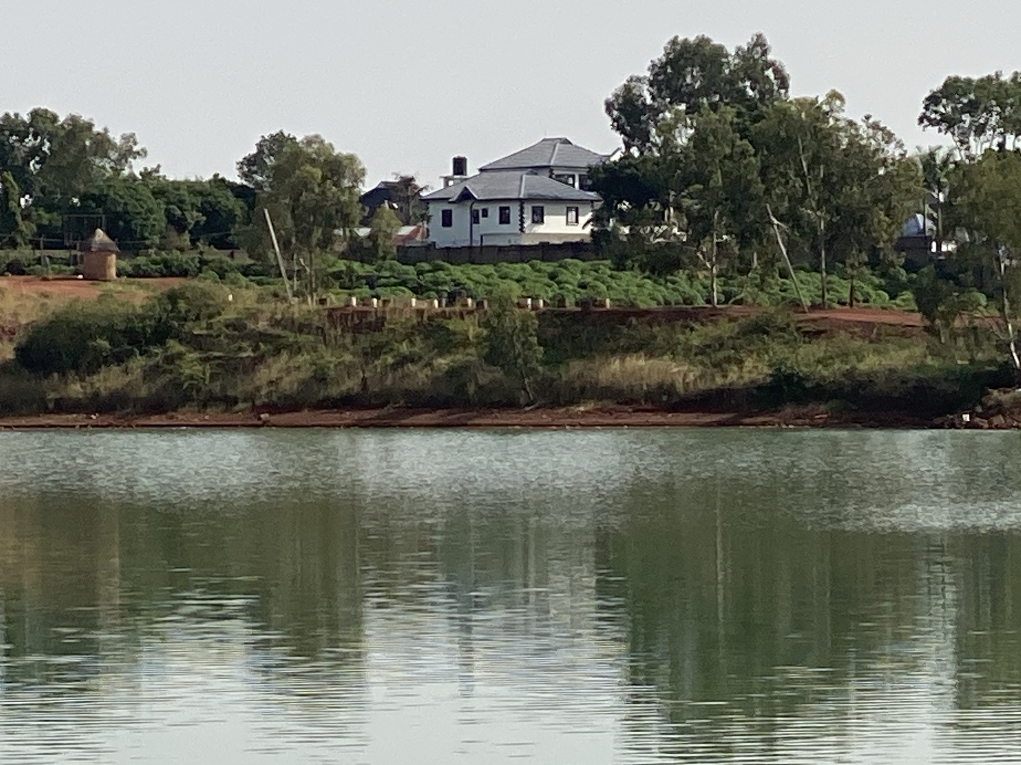 A wide shot captures a scene dominated by a body of water, reflecting the surrounding landscape on its slightly rippled surface. Above the water, a reddish-brown embankment is topped with vegetation, creating a natural barrier. On the embankment, there's a small, round, hut-like structure to the left. A two-story white building with a grey roof, possibly a house, stands further in the background. Trees, predominantly green, are dispersed throughout the scene, framing the buildings and the water. The sky is overcast.
Location: Unknown, Unknown.