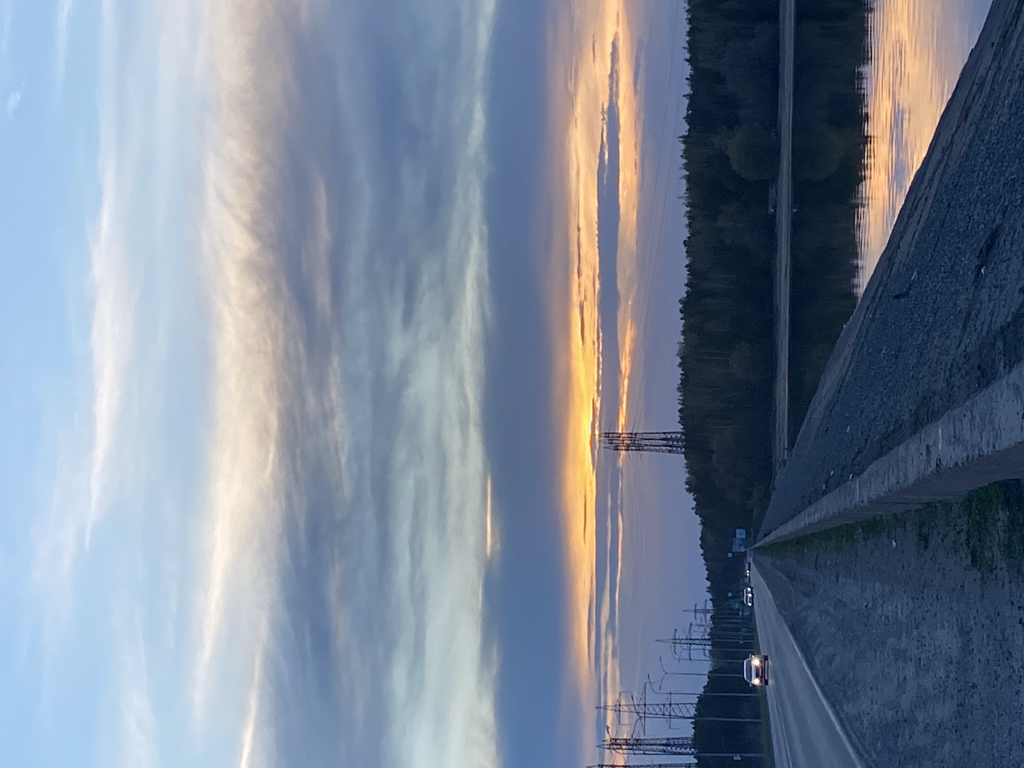 The image shows a road in the city of Noviy, Russia at dusk. Power lines run along the side of the road. A car is traveling on the road. The sky has clouds reflecting the colors of the sunset. The scene is calm and quiet.