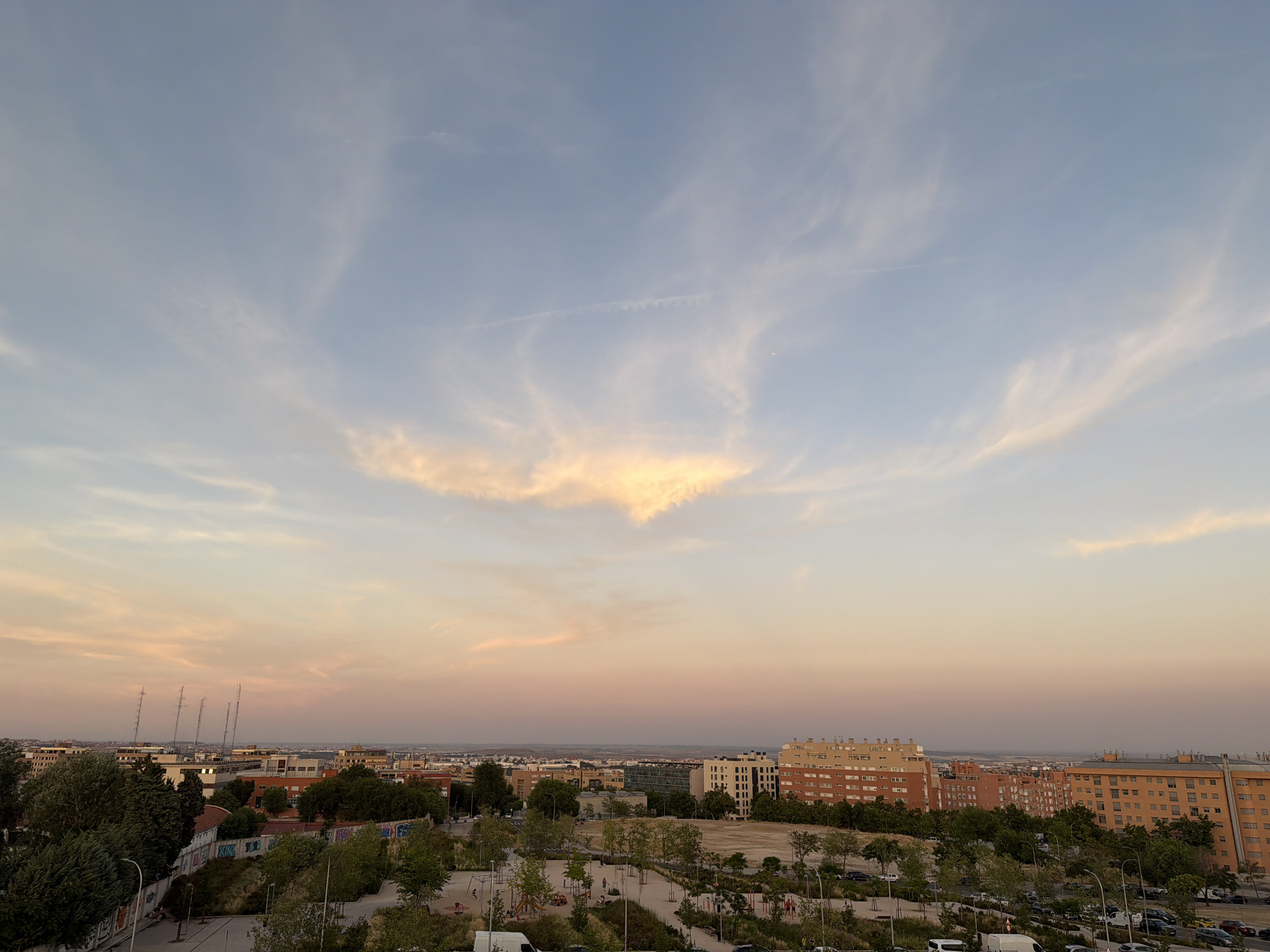 The image captures a cityscape at dusk, with the sky displaying hues of blue, pink, and orange, indicative of sunset or sunrise. Wispy clouds are scattered across the sky, with a more prominent, illuminated cloud formation near the center. Below the sky, the urban landscape unfolds with buildings of varying architectural styles and colors, predominantly residential blocks. Green trees and parks dot the foreground and middle ground, suggesting a city with integrated green spaces. There are signs of infrastructure, including streetlights, a road, and parked vehicles. The overall impression is one of a peaceful urban environment during the transition between day and night. The image appears to be taken from an elevated vantage point, offering a panoramic view of the city.