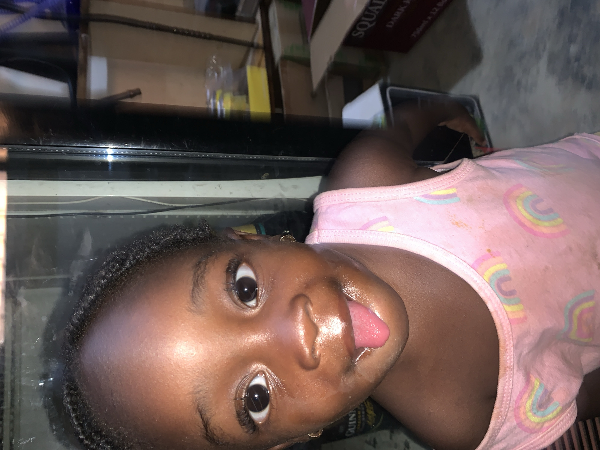 A young African girl with dark skin, wearing a pink shirt with rainbow designs, lies on her side with her tongue sticking out. She appears to be on or near a black countertop. The background includes various boxes and items, suggesting an indoor environment in an unkempt setting. The location, Kabong, Nigeria, is specified.