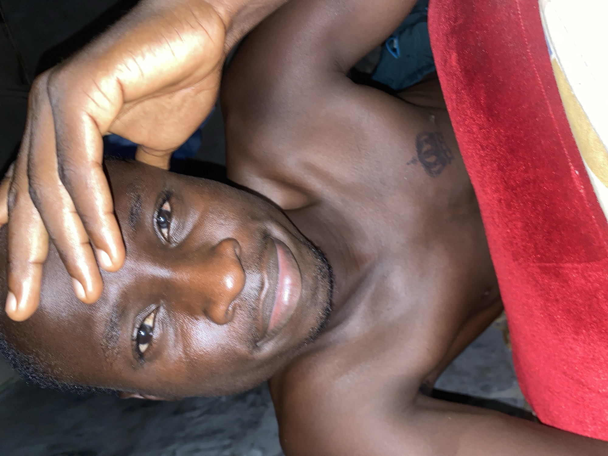 The image shows a shirtless man with dark skin lying down. He has short black hair, dark eyes, and a slight beard. He is holding his right hand near his forehead. There is a small tattoo of a crown on his chest. A red cushion is on the right side of the image. The location is reported to be Narayi, Nigeria, but the interior setting provides no specific environmental details to confirm this.