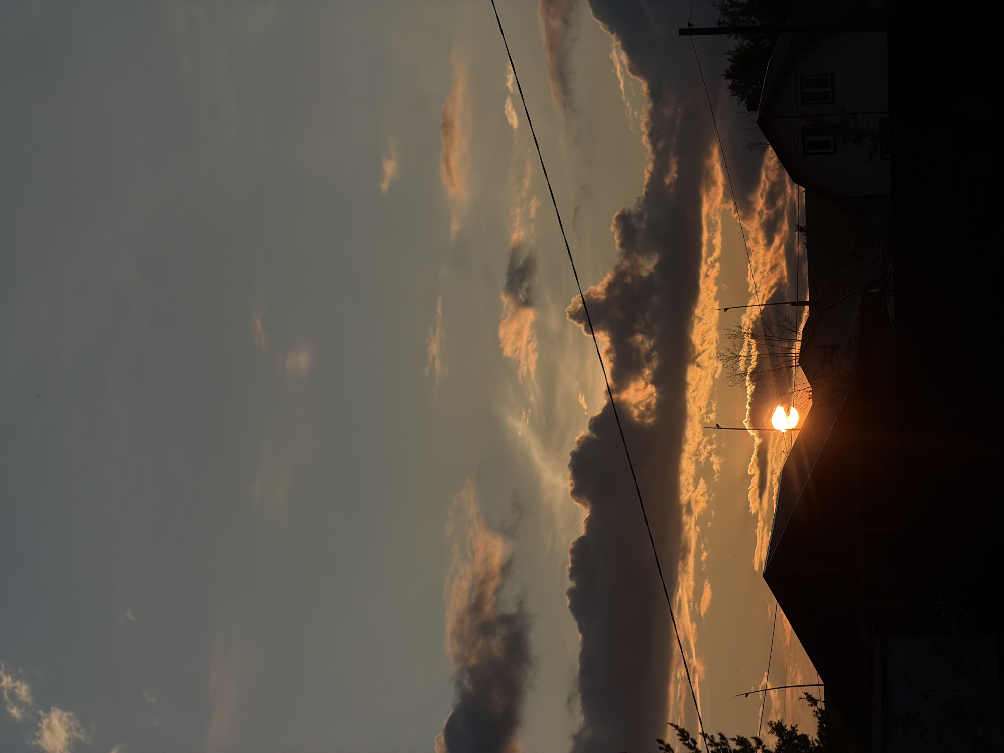 The image depicts a sunset over a residential area in Emel'yanovo, Russia. The sun, a bright orb of orange light, is partially obscured by dark, dramatic clouds, casting warm hues across the sky. Silhouetted against the sunset are the dark roofs of several houses, with the sharp peak of one prominent roof in the foreground. Electrical wires stretch horizontally across the frame, dividing the sky and adding a man-made element to the natural scene. The sky itself is a gradient of soft blues and oranges, with lighter clouds catching the last rays of sunlight. The overall impression is one of a peaceful, albeit dramatic, end to the day in a rural or suburban setting.