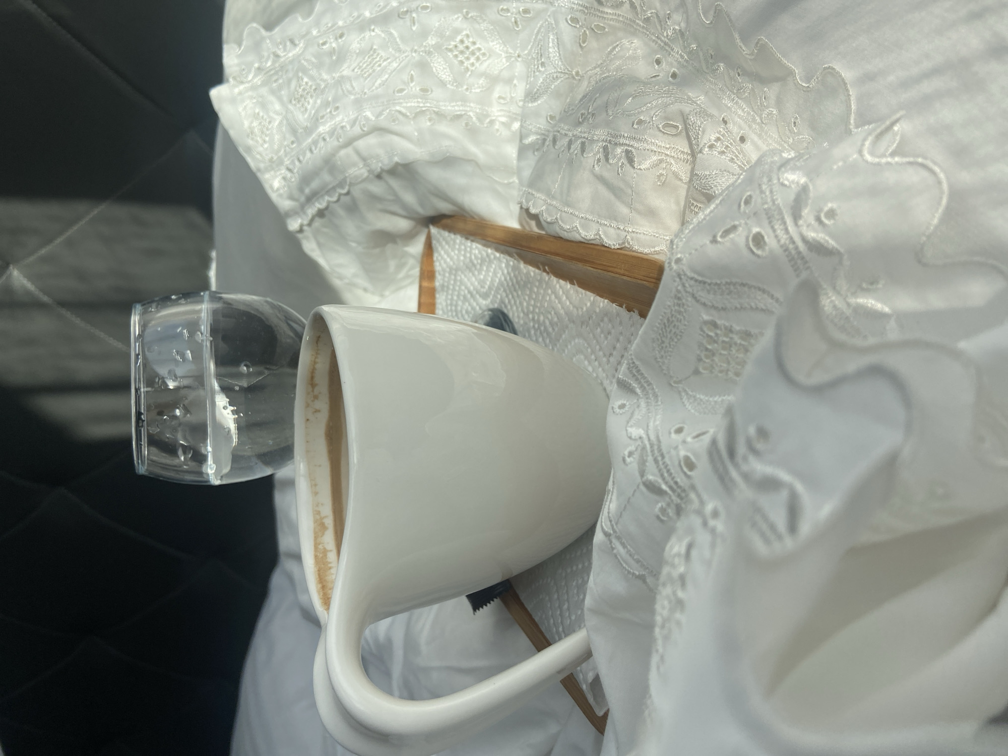 The image features a white ceramic mug with a coffee-colored liquid inside and a clear glass filled with water, both resting on a light brown wooden tray. The tray is placed on what appears to be a bed covered with white, intricately embroidered linens. The background includes a dark, quilted or patterned surface. The location is not identifiable as Budva, Montenegro from the provided information.