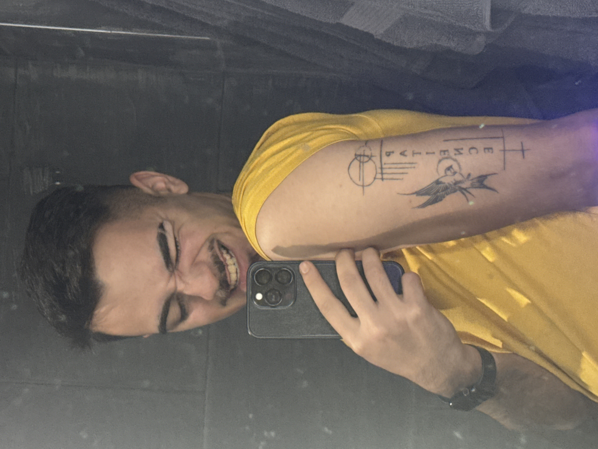 The image shows a man taking a selfie in what appears to be a restroom, given the mirror backdrop. He is wearing a yellow t-shirt and has a dark wristband or watch on his left wrist. He's holding a smartphone with three prominent camera lenses in his right hand. His right arm is flexed, showcasing a tattoo featuring geometric shapes, the word "PATIENCE" aligned vertically, and a stylized bird. His teeth are showing in a grimace. The background is a dark, possibly tiled, wall, slightly obscured by the mirror's reflection and possible condensation. There is no visual information to determine if the subject is in Sofia, Bulgaria or any other location.