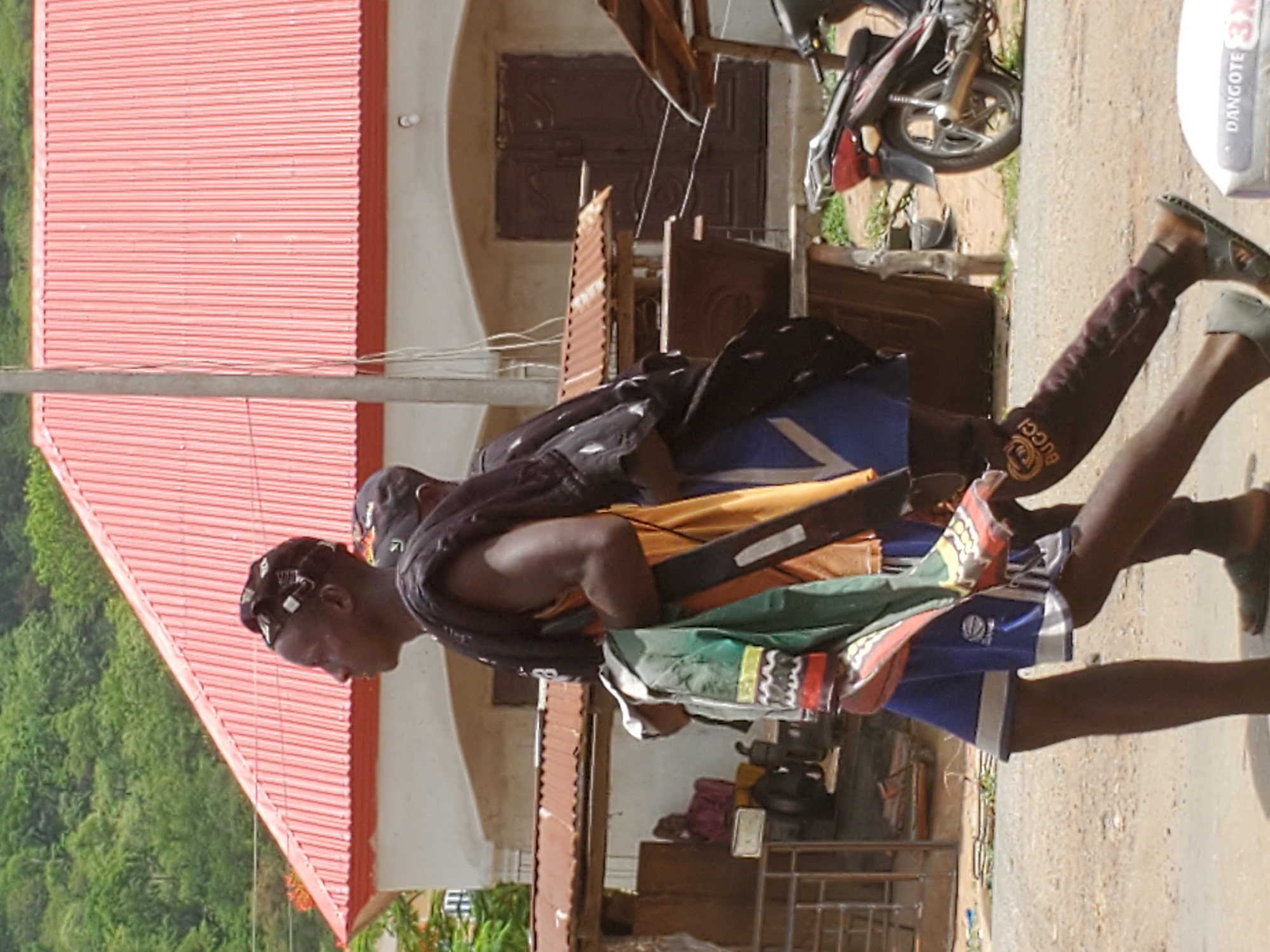 The image shows a man in Mararaba, Nigeria, wearing traditional attire. He has a dark complexion, a shaved head with decorative elements, and is wearing a black shawl over his shoulders. The man is carrying a bundle of colorful fabrics. He's wearing shorts with a blue and white pattern. The background features a red corrugated iron roof of a building, a motorcycle parked nearby, and a sack labeled "Dangote". The environment appears to be a residential area. The ground is a concrete surface. Another person is partially visible behind the main subject.