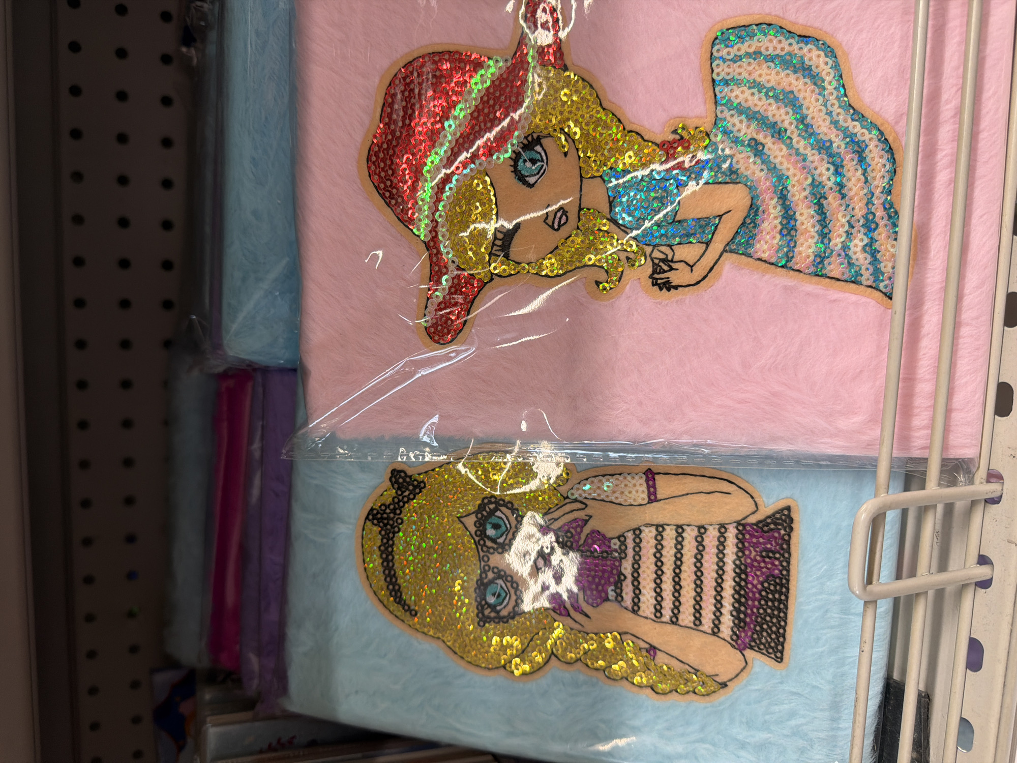 The image shows two fluffy notebooks with sequined doll appliques, displayed on a store shelf. The notebook on the left is light blue and features a doll with blonde curly hair, glasses, and a striped dress. The notebook on the right is pink and features a doll wearing a red sequined hat and a blue and white striped dress. The notebooks are partially wrapped in plastic. Behind the notebooks, stacks of other colorful notebooks and stationery items are visible on the shelf. The shelf itself is a metal rack with horizontal bars. The image appears to be taken in a retail setting, possibly a stationery or toy store. The specific location in Pazardzhik, Bulgaria is not discernable from the image.