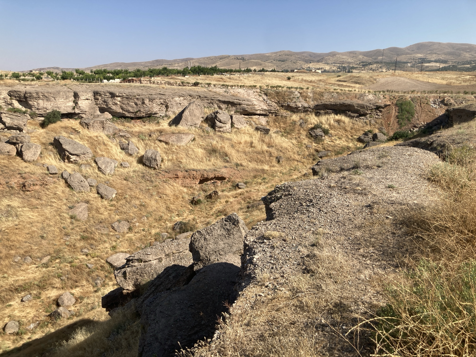 The image captures a dry, rocky landscape in Avanos İlçe Merkezi, Turkey, under a clear blue sky. In the foreground, a steep embankment of gravelly soil and dry grass slopes downwards. Beyond this, a deep, dry gorge dominates the scene, with steep, eroded rock faces and scattered boulders. The gorge floor and surrounding slopes are covered in dry, yellowed grass, indicating an arid climate. In the middle ground, a prominent, layered rock formation forms the edge of the gorge. Further in the distance, the terrain transitions to rolling hills dotted with sparse vegetation and some cultivated fields. A line of trees is visible on a plateau in the far distance, with some structures and electrical poles scattered across the landscape. The overall impression is one of a rugged, semi-arid environment.