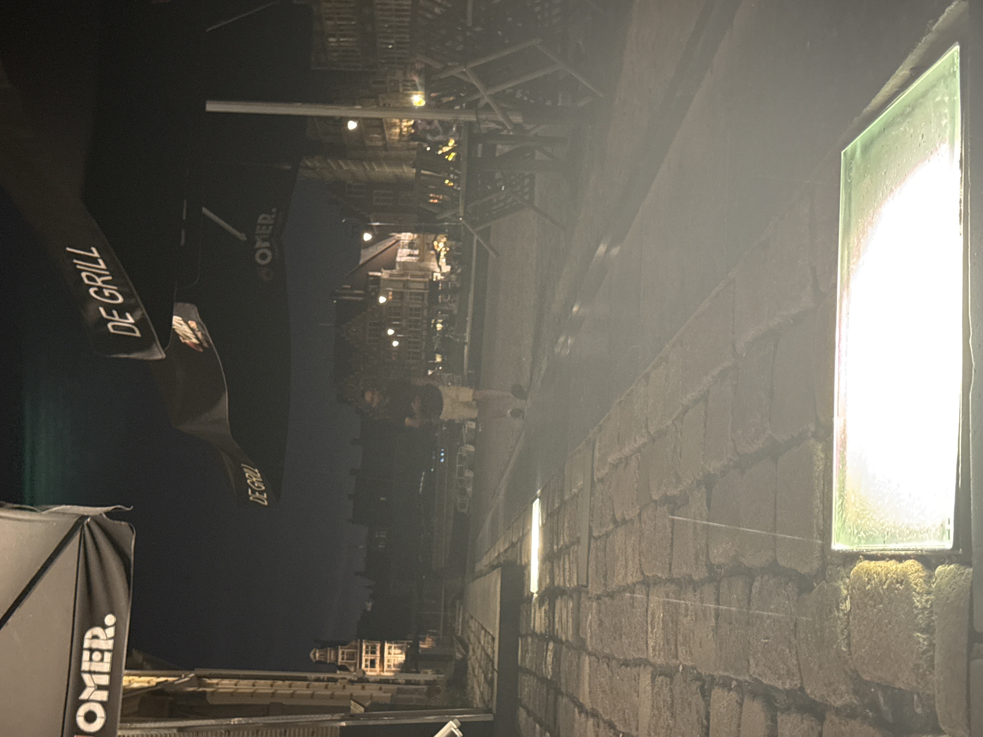 This is a night-time photograph taken outdoors on a cobblestone street in Ghent, Belgium, likely in a public square or near a canal. In the foreground, a horizontal, illuminated rectangular light fixture is set into the cobblestones. The street leads back to a canal or body of water with boats docked along its edge. Across the water, there are buildings with illuminated windows, suggesting a lively area with restaurants or businesses.
In the middle ground, a person is walking away from the camera. They are wearing light-colored shorts and a dark t-shirt. The street is lined with outdoor seating areas, including tables and chairs, some of which are stacked or overturned, indicating the end of business hours or preparations for closing. Several large black umbrellas with logos are visible, including one with the text "OMER." and another with "DE GRILL." The overall atmosphere is dimly lit, with streetlights and building lights providing illumination, creating a somewhat hazy and atmospheric scene. The architecture of the buildings suggests a historic European city.