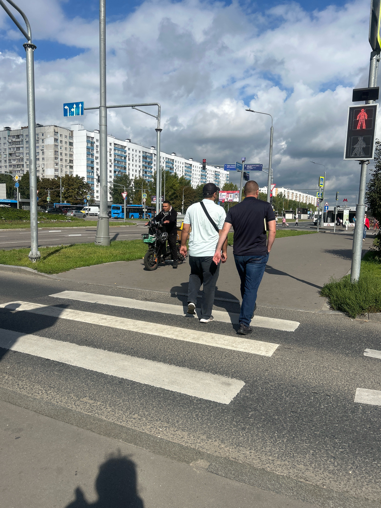 The image depicts a street scene in Moscow, Russia, on a partly cloudy day. Two men are walking across a pedestrian crossing, one in a light blue t-shirt and dark pants, the other in a dark blue t-shirt and blue jeans. A person on a black electric scooter is stopped on the sidewalk behind them. In the background, there are tall apartment buildings, a street with traffic including buses and cars, and traffic signals. The pedestrian signal displays a red walking figure, indicating that pedestrians should not cross. The scene appears to be a typical urban environment with residential and commercial elements.