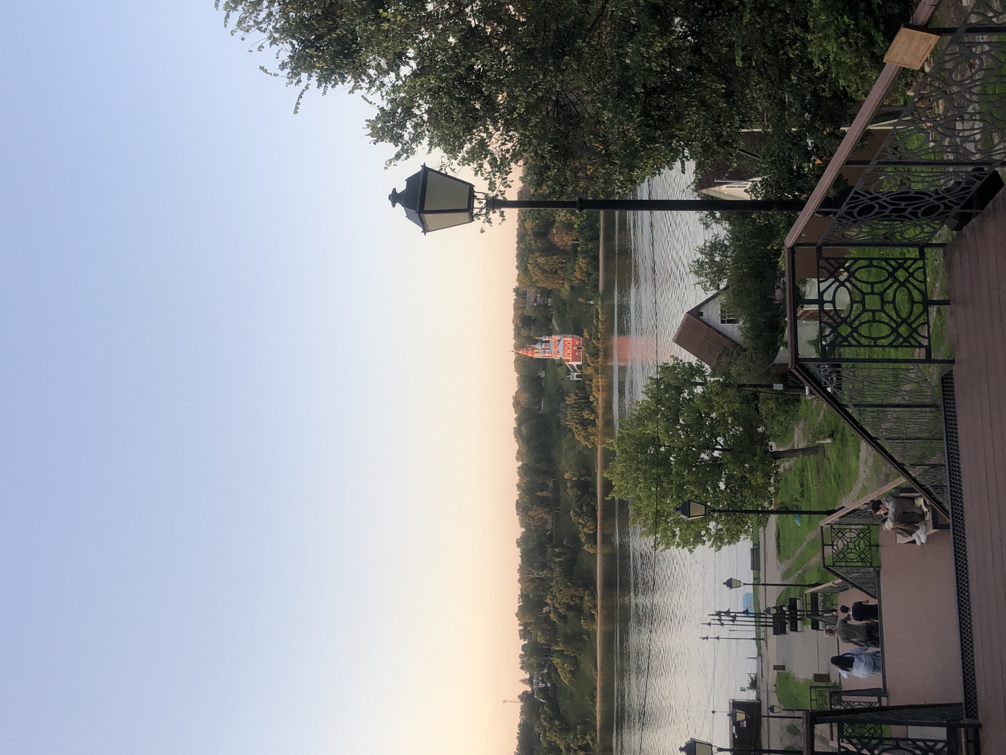 The image depicts a scenic view of the Volga River at dusk in Tutaev, Russia. In the foreground, a wooden staircase with ornate metal railings leads down towards a promenade along the river. Several people are visible on the stairs and promenade, some sitting and others walking. A vintage street lamp stands prominently on the right side of the frame. Across the river, a lush green hillside is dotted with trees and houses. A striking red and white church with multiple spires stands out on the hillside, its reflection shimmering in the water. The sky is a soft gradient of pale blue and pink, indicating the end of the day.
