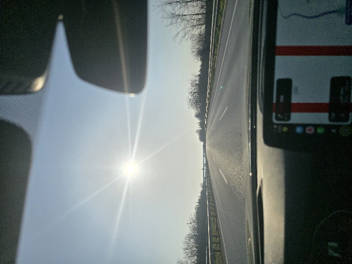 The image displays a forward-facing view from inside a vehicle traveling on a multi-lane highway in Babaeski, Turkey. The bright sun, visible in the upper-left quadrant of a clear blue sky, casts strong light onto the scene, causing a starburst lens flare. The asphalt highway extends into the distance, marked by white dashed lines and bordered by metal guardrails on both sides. Beyond the guardrails, deciduous trees with bare branches line the road. An overpass or bridge structure is discernible in the far distance. The vehicle's dark dashboard occupies the lower portion of the frame, featuring a central infotainment screen on the right that displays a map interface with two distinct vertical red lines. No individuals are visible.