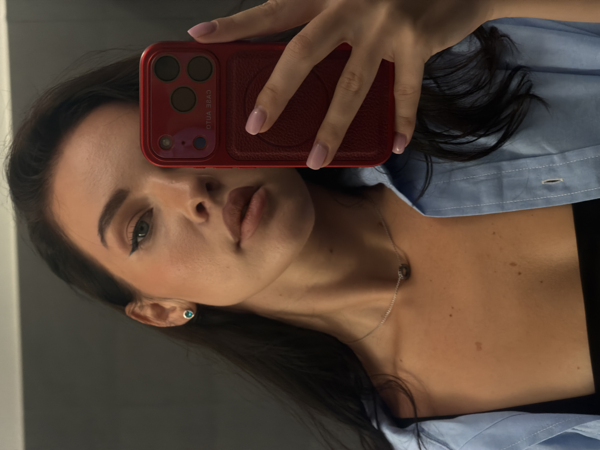 A woman is taking a selfie with a red phone, the camera obscuring part of her face. She has dark hair, fair skin, and is wearing a light blue button-down shirt. Her nails are painted a light pink color. She is also wearing a silver necklace. The background appears to be a gray wall. The location is not evident from the image.