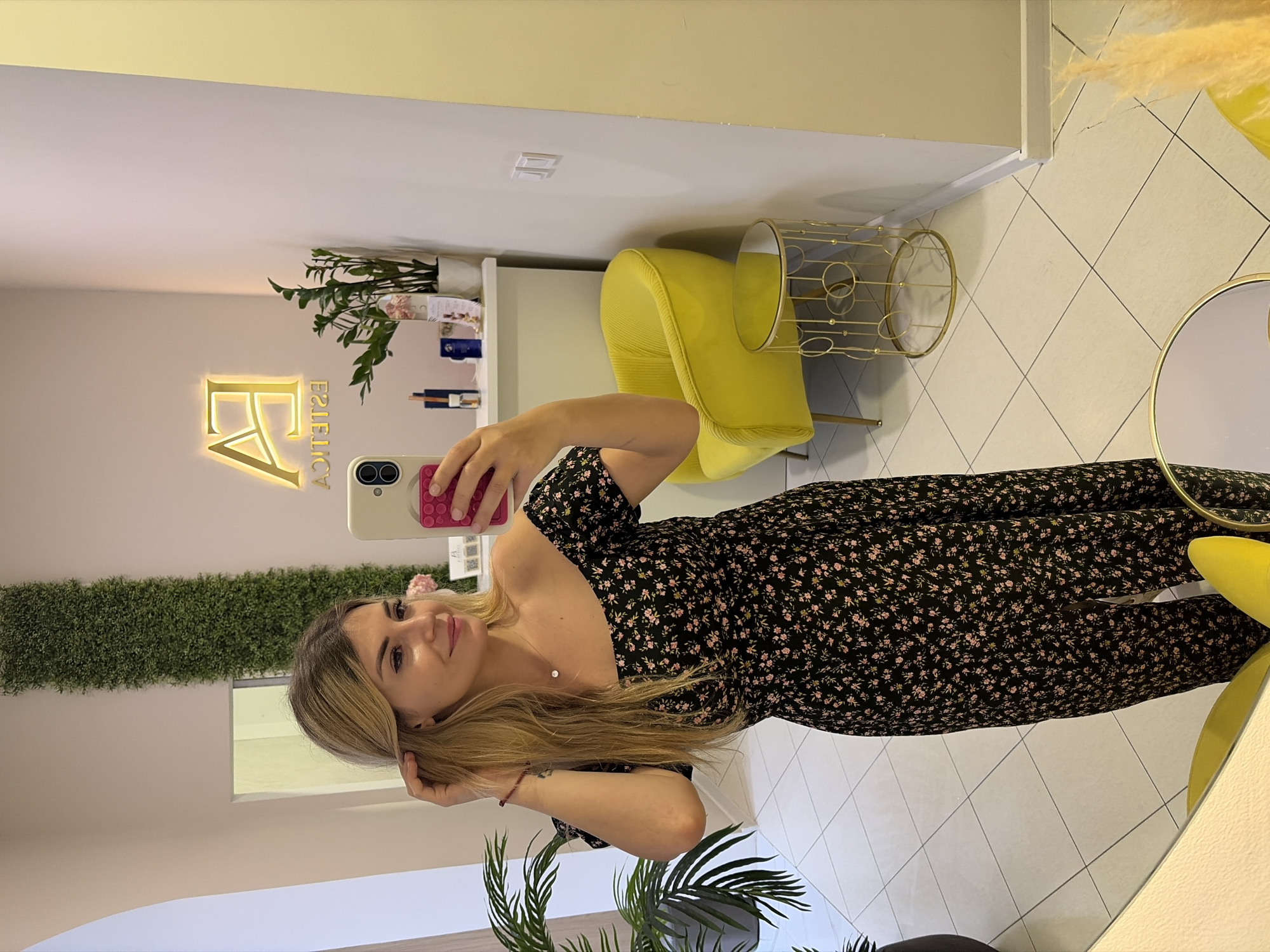 A young woman with blonde hair, wearing a black off-the-shoulder dress with a floral print, is taking a mirror selfie in what appears to be a beauty salon or spa in Budva, Montenegro. The salon has modern decor with a yellow accent chair, a gold circular side table, and plants. A neon "EA ESTETICA" sign is visible on the wall. The woman is holding her phone in her right hand and touching her hair with her left. The floor is tiled.