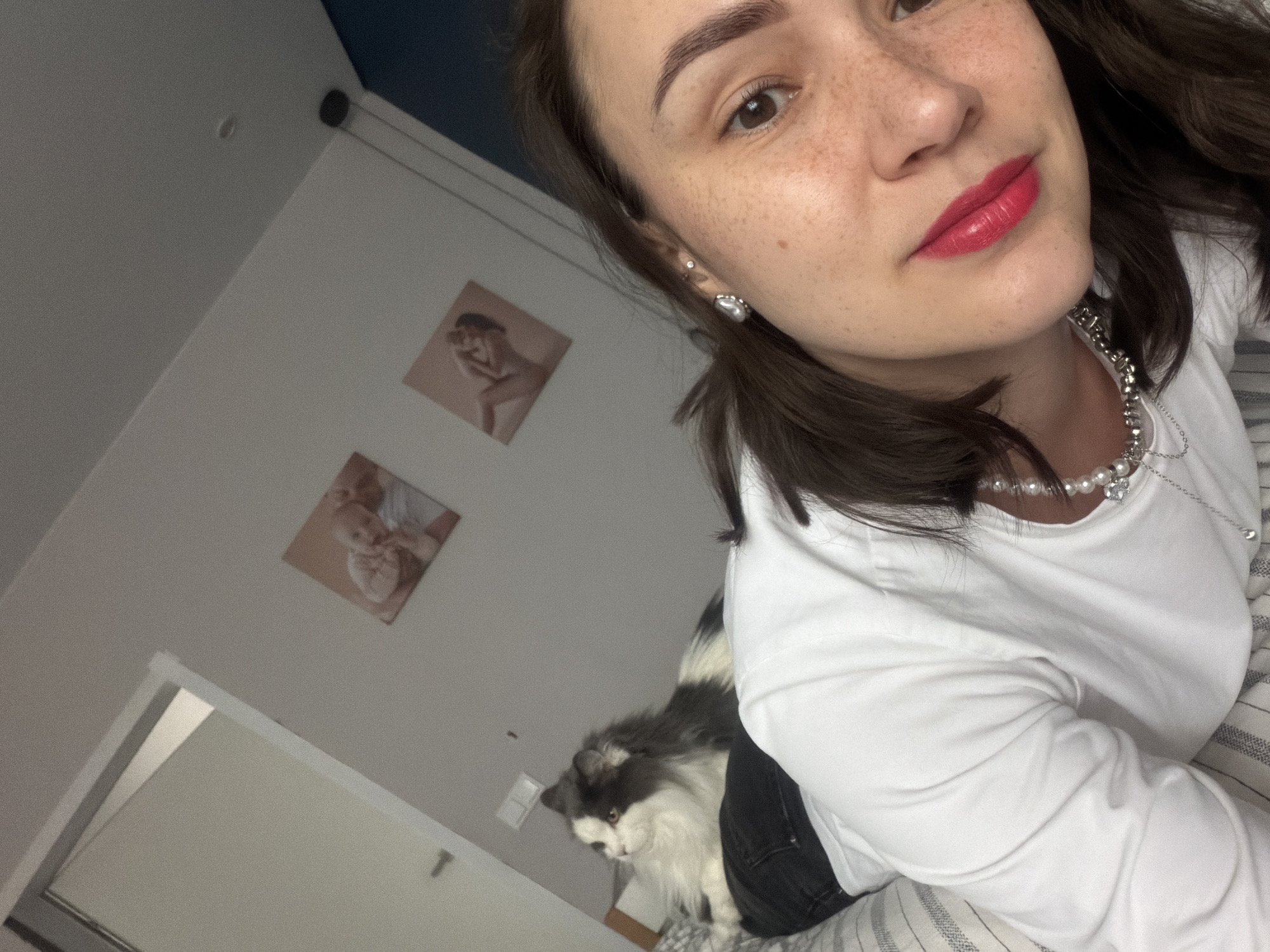 A selfie shows a young woman with brown hair and fair skin, wearing red lipstick and a white long-sleeved shirt with a pearl necklace. She has freckles on her face. In the background, two framed pictures hang on a light-colored wall. A gray and white cat is visible near the woman's legs. The location is presumed to be a residential interior in the city of Chaikovsky, Russia.