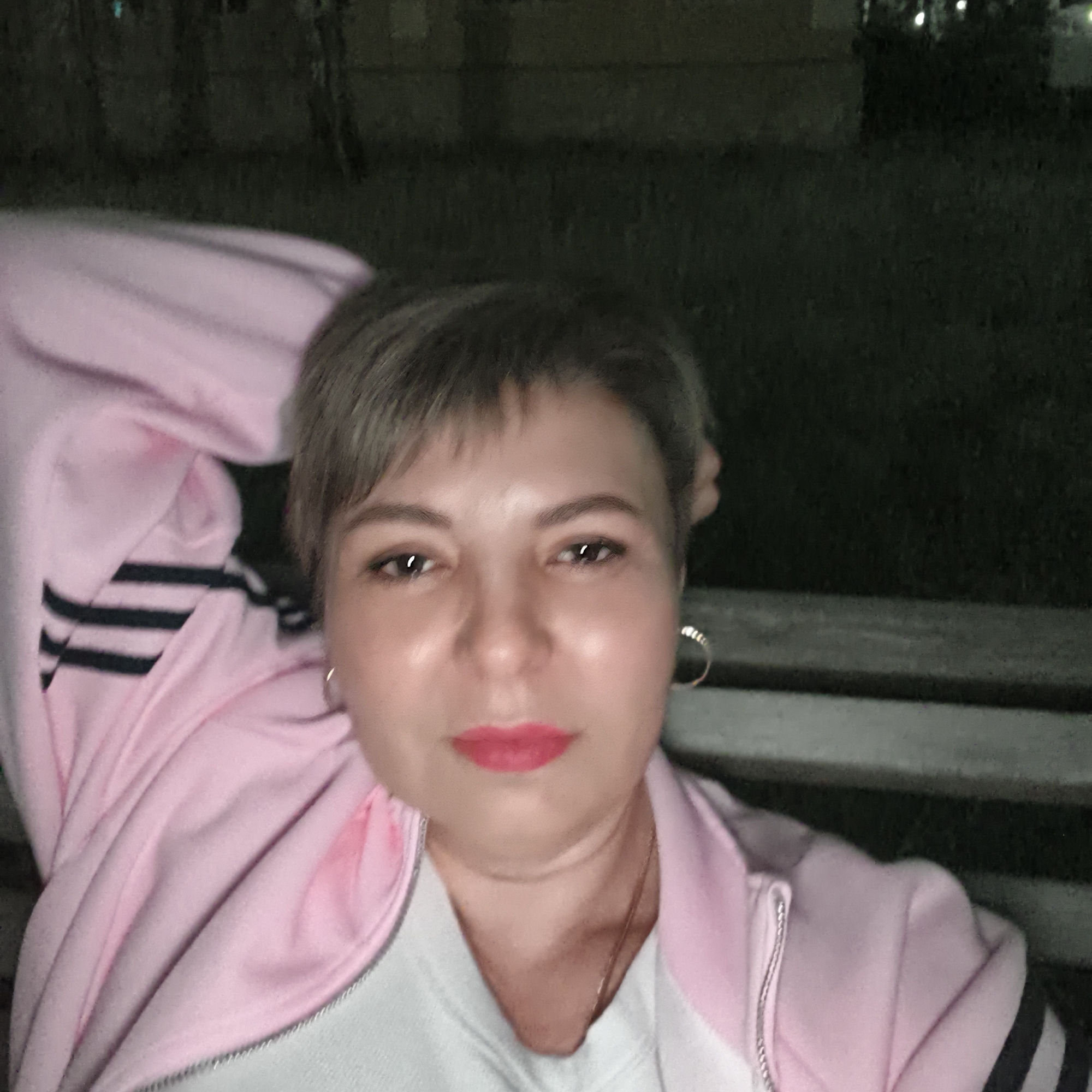 A medium shot of a fair-skinned woman with short, graying hair, red lipstick, and wearing silver hoop earrings. She is wearing a pink athletic jacket with three black stripes on the sleeves over a white shirt. She is reclining on a wooden bench outside at night. The background is dark and indistinct, but appears to be a grassy area with some trees and buildings visible. The location is Варзи-Ятчи, Russia.