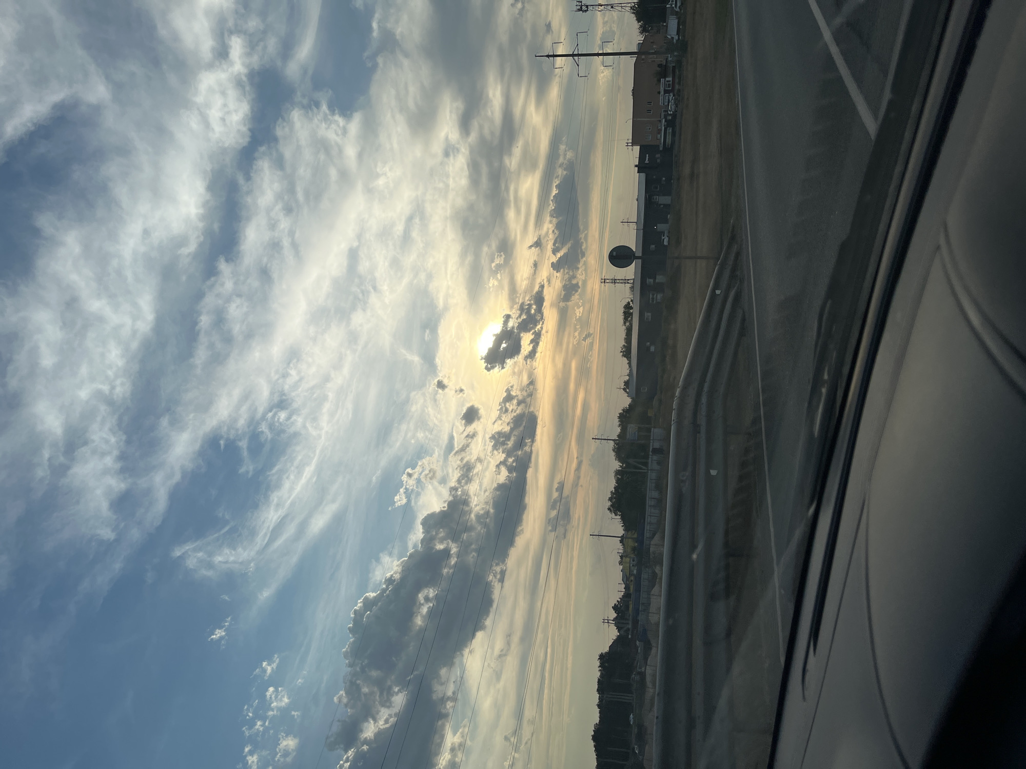 The image depicts a view from inside a vehicle looking out at a landscape under a dramatic sky. The sky is a prominent feature, filled with a mix of blue sky and various clouds, including wispy cirrus clouds and darker, more substantial formations. The sun is partially obscured by clouds, creating a soft, diffused light that casts a warm glow across the scene, suggesting it might be dawn or dusk.
In the middle ground, there is a road with visible lane markings. Beyond the road, the terrain appears somewhat dry and grassy. Several industrial or commercial buildings are visible in the distance, characterized by their rectangular shapes and varied rooflines. Power lines and utility poles are scattered throughout the landscape, indicating the presence of infrastructure. Some trees and vegetation can be seen lining the horizon.
The foreground shows the interior of the vehicle, specifically the dashboard and windshield, which are slightly reflective, capturing a distorted view of the outside world. The perspective suggests the vehicle is moving or stationary by the side of a road.
The location is specified as Shumeika, Russia. This suggests the landscape and buildings might be characteristic of the Russian countryside or an industrial area within that region. There are no people visible in the image. The overall impression is one of a transitional landscape, perhaps on the outskirts of a town or city, with a focus on the natural elements of the sky and the man-made elements of infrastructure and buildings.