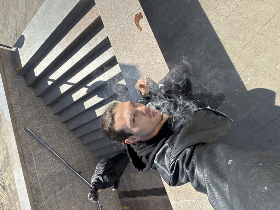A high-angle selfie captures a male subject exhaling cigarette smoke in a public stairwell in Sofia, Bulgaria, under bright daylight. The subject, positioned in the upper portion of the frame, has dark hair pulled back, a trimmed beard, and is wearing a black leather jacket over a black hooded sweatshirt. He holds a lit cigarette in his left hand, with a visible plume of smoke emanating from his mouth and around his face as he looks directly into the camera. In the background, below and to the left of the main subject, another individual dressed in a dark coat and carrying a backpack walks down the concrete stairs. A metal handrail runs alongside the stairs. Strong sunlight creates prominent diagonal shadows across the steps and the grey tiled ground level. A reddish-brown, irregular stain is visible on the tiled ground to the right of the smoking individual.