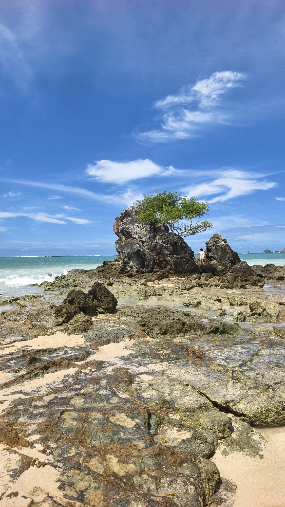 The image depicts a rocky beach scene in Kuta, Indonesia. In the midground, a large, jagged rock formation rises from the beach, topped with a green tree. A single person, dressed in light clothing, is visible on the rocks. The foreground is filled with smaller rocks and sand, some of which appear wet. The ocean is visible in the background, with gentle waves breaking near the shore. Above, the sky is bright blue with scattered white clouds.