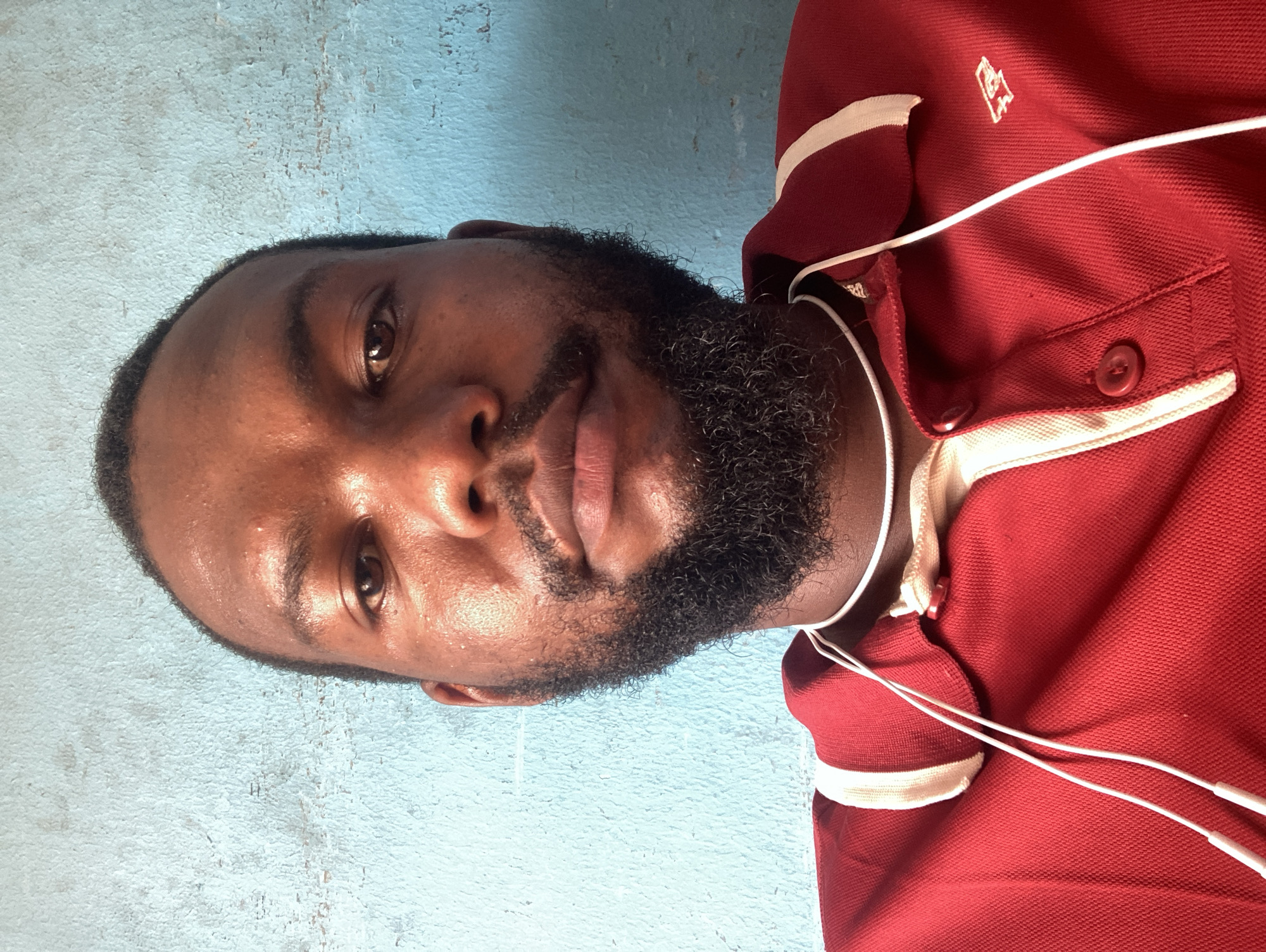A portrait of a man with dark skin and a beard wearing a red polo shirt with white trim. He is wearing white earphones. The background appears to be a light blue wall. It is not possible to determine the location in Jos, Nigeria from the image.