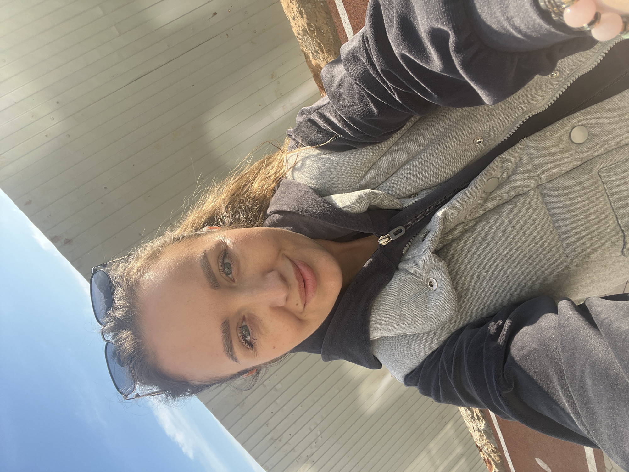 A woman is taking a selfie outdoors. She has brown hair, green eyes, and is wearing sunglasses atop her head. She is wearing a grey jacket with a black hoodie underneath. The background includes a clear blue sky, a white painted wooden structure, and brickwork. The location appears to be somewhere with architecture that could be found in Pervomaysky, Russia, but without further landmarks, it's difficult to ascertain a specific location.