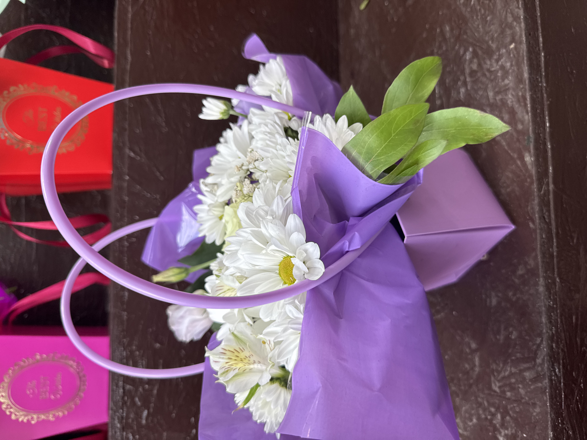 A bouquet of white daisies and alstroemerias is presented in a small, square, light purple gift box. The flowers are wrapped in purple tissue paper and have light purple handles on the gift box. The bouquet is placed on a dark brown, textured surface. In the background, two gift boxes are visible, one pink and one red, both decorated with gold circular embellishments. The scene appears to be indoors, possibly in a flower shop or a retail setting in the city of Kamianske, Ukraine.