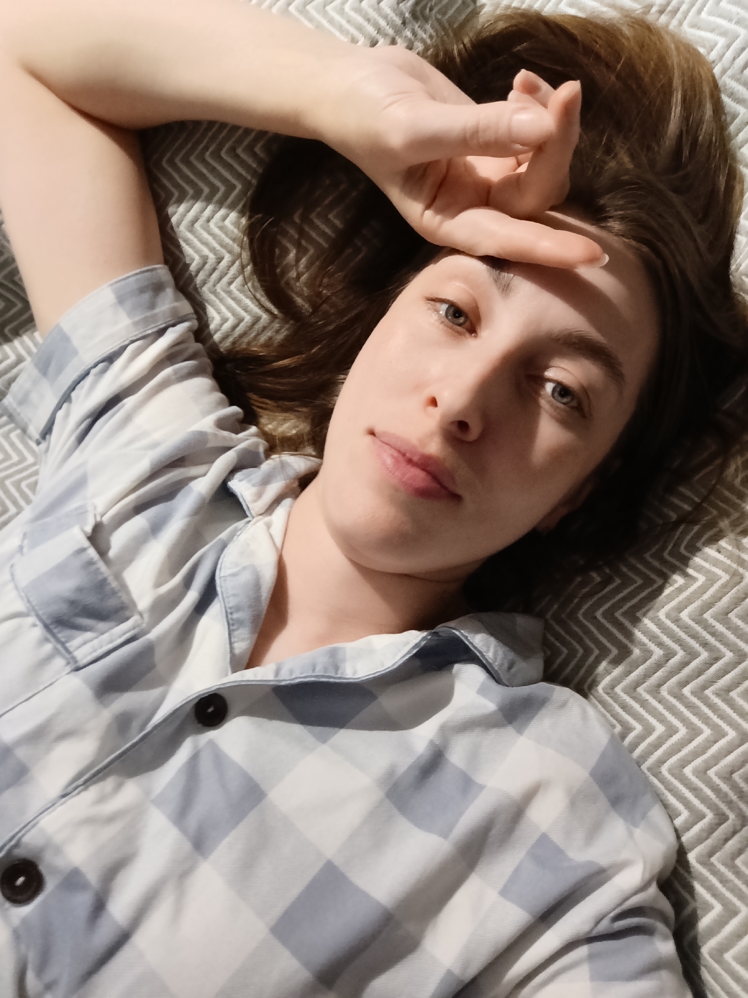 A young woman with light skin and shoulder-length brown hair lies on a gray and white chevron-patterned surface, likely a bed. She is wearing a blue and white checkered button-down shirt with a collar. Her right arm is extended overhead, with her hand resting on her forehead. She is looking directly at the camera with a neutral expression. The background provides no information about the location in Chaikovsky, Russia.