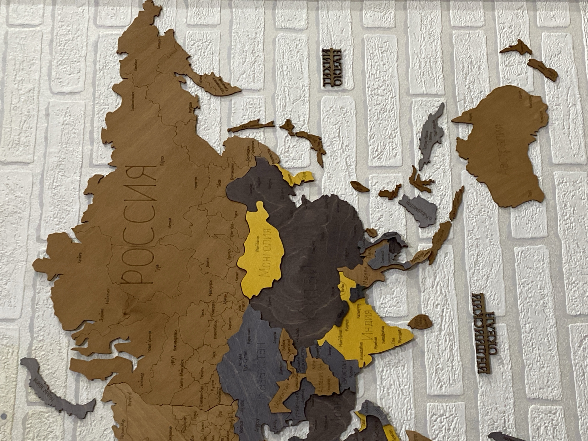 The image shows a wooden world map mounted on a white brick wall. The map is intricately detailed, with borders of continents and countries delineated and names of countries and cities etched onto them in Russian. Some countries are colored differently, such as India and Mongolia, which are yellow. The map is a decorative piece, likely in a home or public space. There is no discernible location in the city of Марковский, Russia, visible in the image.