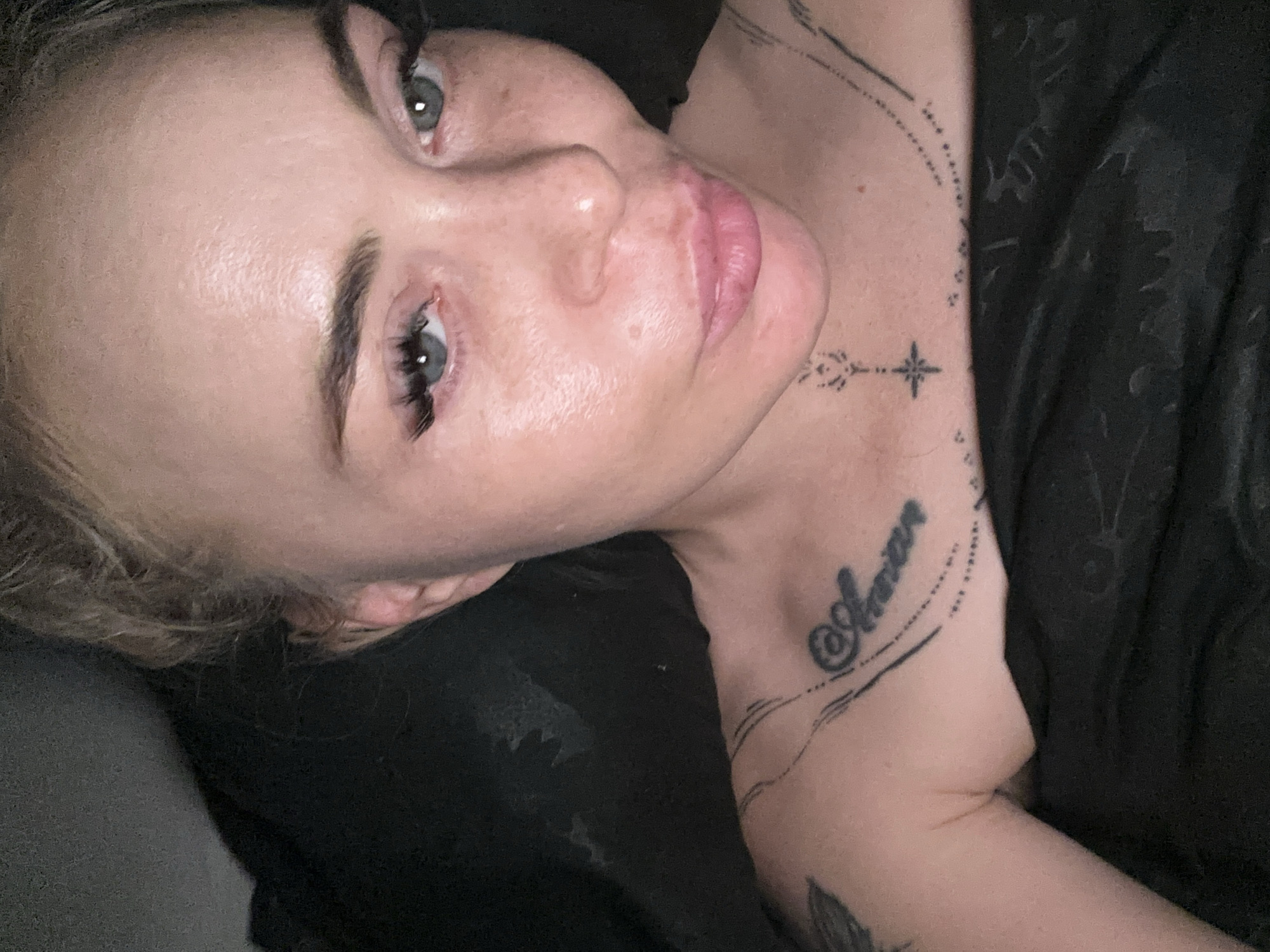 A close-up, high-angle perspective captures a light-skinned individual, likely female, reclining in a dimly lit indoor setting. The individual has light, wavy hair, partially pulled back, with visible strands framing the face. Their eyes are light blue-grey, looking directly at the camera, and feature prominent, dark eyelashes, notably voluminous on the left eye. The eyebrows are dark and well-defined. The facial skin and visible upper chest are fair, showing some natural texture, minor blemishes, and a slight sheen. The lips are full, light pink, and slightly parted, forming a subtle pout.
Several tattoos are visible on the upper torso:
* On the left clavicle/shoulder, a script tattoo reads "@Amore" (or similar), encased within decorative dotted lines.
* Centered at the base of the neck, a small, stylized asterisk or four-pointed star-like symbol is present, accompanied by geometric lines extending upwards towards the chin.
* A fragment of a larger, dark tattoo, possibly organic or floral in design, is visible on the upper left arm/shoulder.
* Additional faint dotted lines trace along the collarbones.
The individual is partially covered by a dark, possibly black, fabric item, which appears to be a blanket or piece of clothing. The background is uniformly dark and indistinct, suggesting an enclosed space with limited visibility. Lighting illuminates the face primarily from the front, creating soft shadows. The location is in the city of Unknown, Unknown.