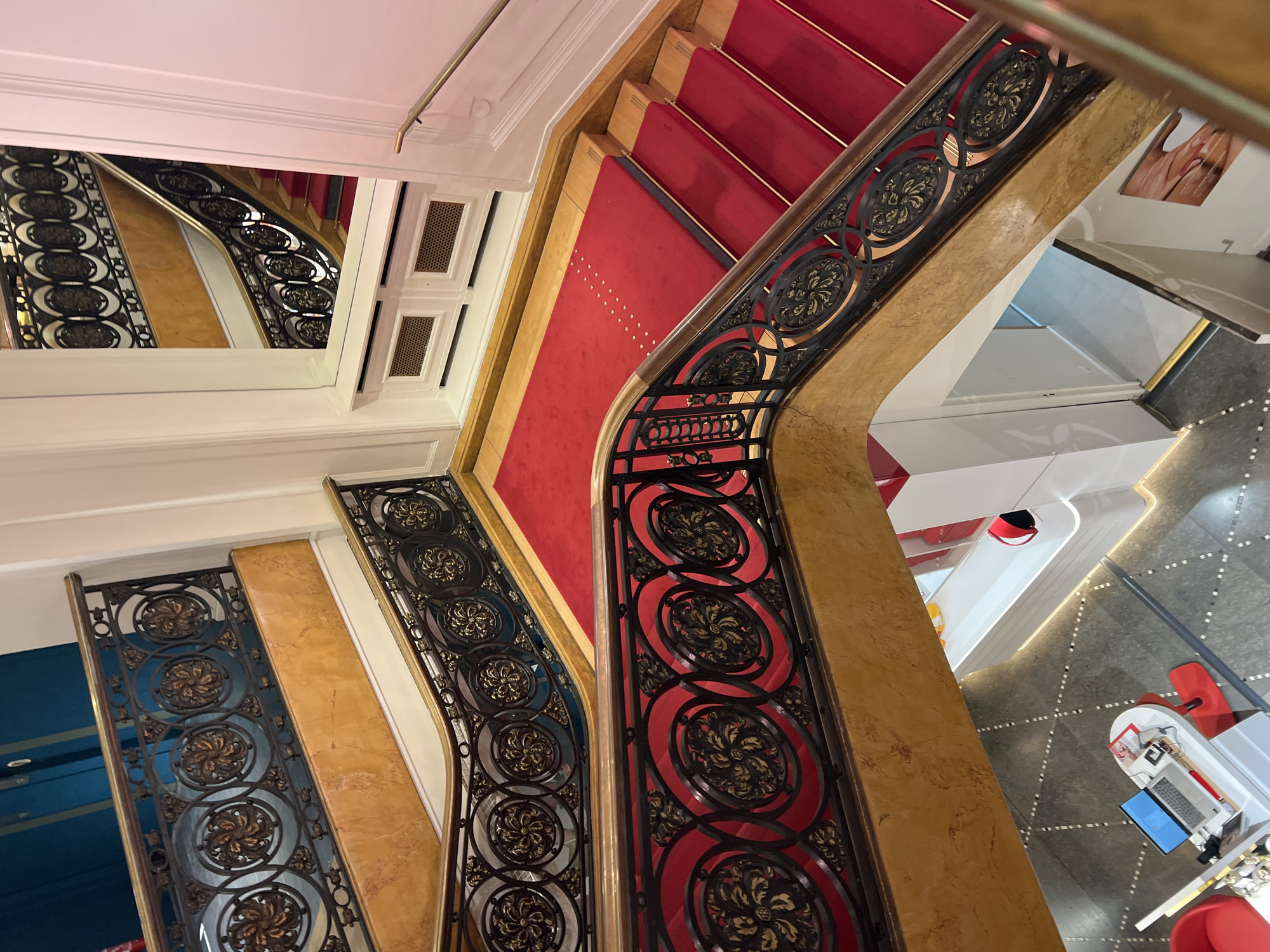 The image shows a grand staircase with ornate black metal railings featuring circular floral designs and a golden marble banister. Red carpet runners with golden accent stripes cover the steps. The staircase winds upwards, and a reflection of the staircase is visible in a large mirror on the wall. Below the mirror, there are two rectangular ventilation grates. The floor at the bottom of the stairs has a grey tiled pattern. In the lower left of the image, there is a white table with a laptop, some papers, and a red desk accessory, and red chairs are visible. The environment appears to be indoors, possibly a hotel, theater, or upscale establishment, with a sense of luxury and historical architecture.