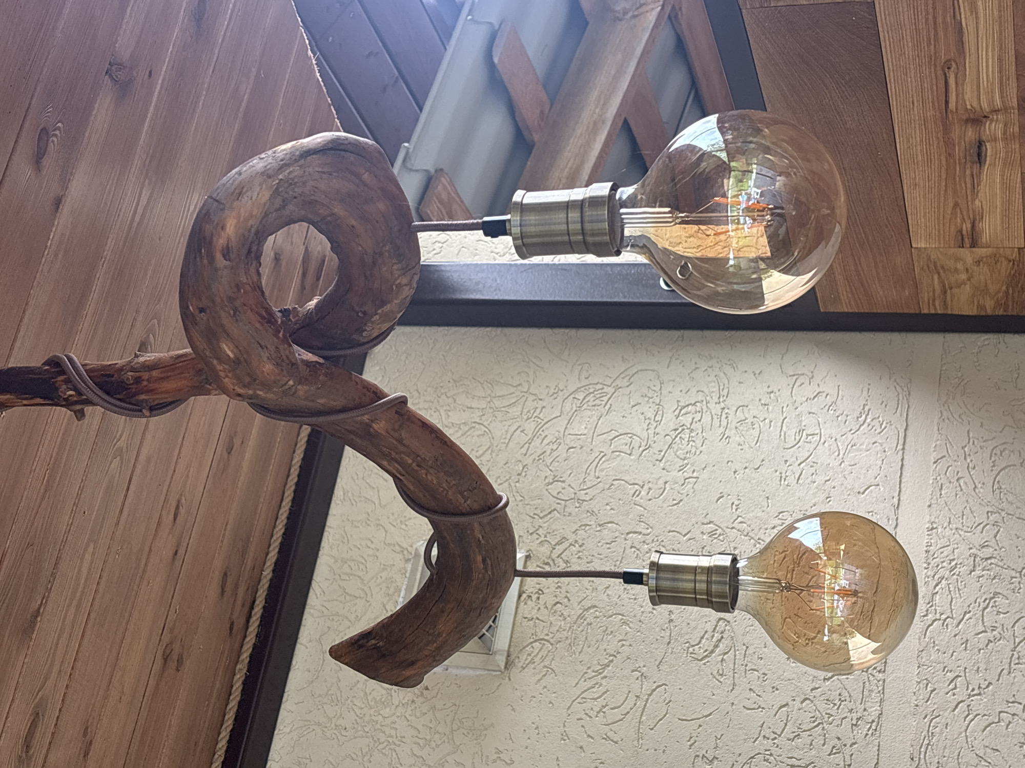 The image shows a lamp fixture. The fixture is constructed from a twisted branch with two light bulbs attached. The light bulbs are vintage-style with visible filaments. The lamp is mounted on a wall with a textured surface. The background includes wooden paneling and other architectural elements. The image appears to be taken indoors. The location is Lahojsk selsaviet, Belarus.