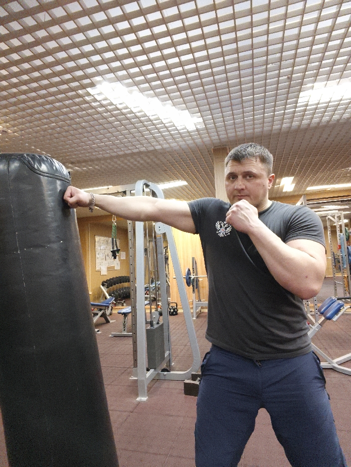 An adult male, identified as a person, is depicted in a gym in Im. Gastello, Russia. The individual, a Caucasian male with short brown hair, stands in a boxing stance, his body oriented slightly towards the left of the frame. He wears a dark grey short-sleeved t-shirt featuring a white emblem on the left chest and dark blue athletic pants. A silver-colored chain bracelet is visible on his left wrist. His left arm is extended, and his left fist is making contact with the upper-middle section of a tall, black cylindrical punching bag positioned to his left. His right arm is bent at the elbow, with his right fist clenched and held defensively near his chin. He maintains eye contact with the viewer.
The gym environment includes a floor covered in dark reddish-brown interlocking rubber mats. In the background, various pieces of gym equipment are visible, including weight machines, benches, dumbbell racks, and kettlebells. The far wall displays light-colored wooden paneling. The ceiling is a white grid-panel design with integrated rectangular fluorescent light fixtures providing illumination to the space.