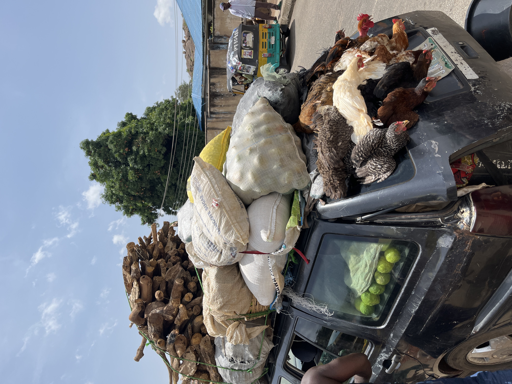 The image depicts a car heavily laden with goods, including sacks of unidentified materials, firewood, and live chickens on the roof. The car is located on a street in Jos, Nigeria, as suggested by the surroundings and typical transportation modes visible. A three-wheeled vehicle (likely a "keke" or auto-rickshaw), a common sight in Nigerian cities, is in the background. A person is walking nearby. The car is packed to capacity, with items also visible inside the vehicle, such as green fruits. The environment shows a typical urban scene with buildings and trees in the background.