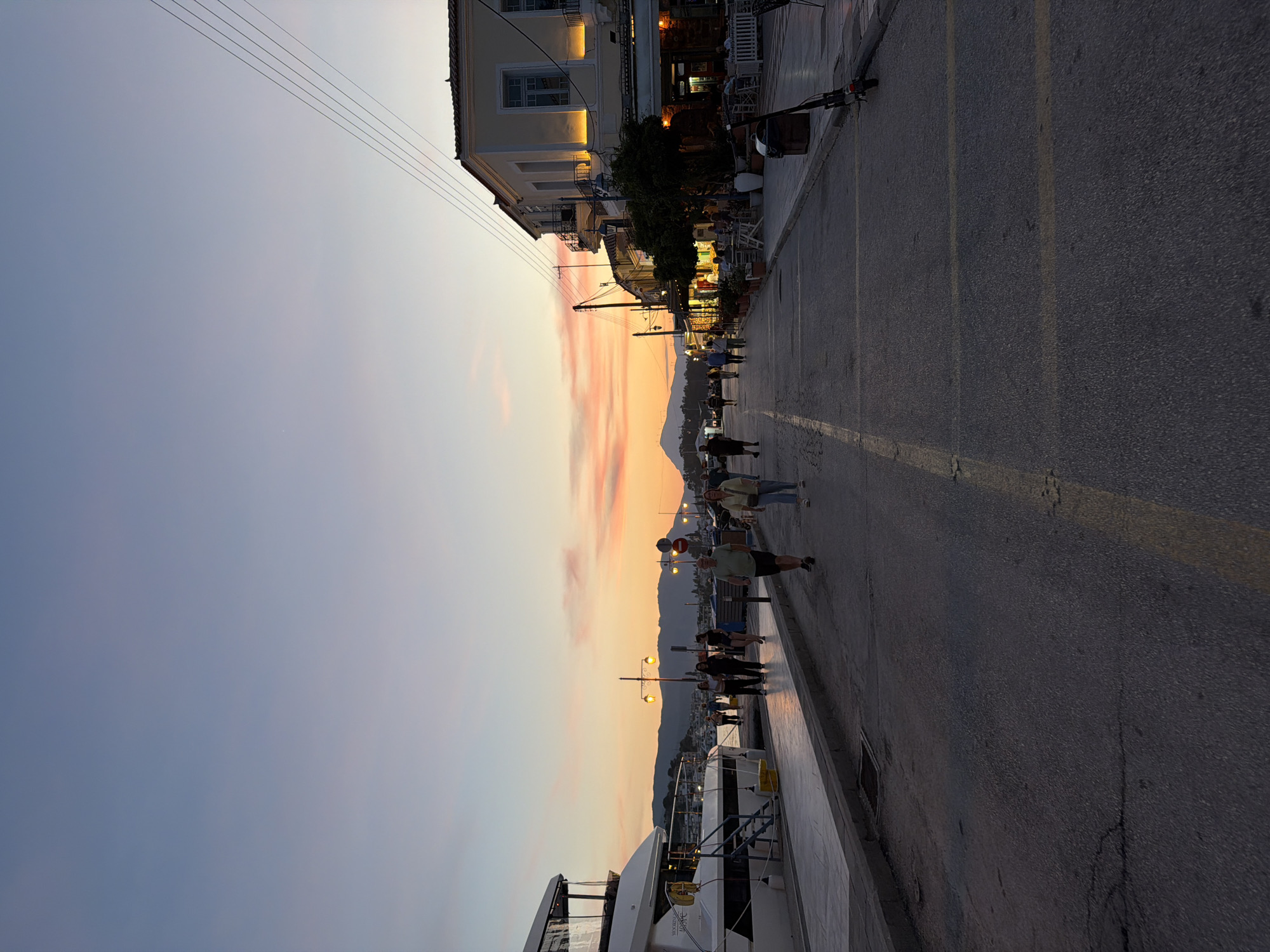 This media file depicts an evening scene along a vibrant waterfront street in a coastal town, identified as Poros, Greece. The time of day is dusk, characterized by a striking sunset illuminating the sky with soft pink, orange, and purple cloud streaks, transitioning to a serene blue overhead.
In the foreground, an asphalt road with yellow lane markings runs parallel to a paved pedestrian promenade. Several people are visible, some strolling casually, others standing, enjoying the evening. A large, white luxury yacht or boat is moored alongside the promenade.
To the right, a row of multi-story buildings with traditional architecture lines the street. These buildings appear to house shops and restaurants, indicated by illuminated windows, awnings, and outdoor seating arrangements. A Greek flag is prominently displayed on one of the buildings. Warm light from streetlights and building interiors casts a soft glow on the scene. A scooter or bicycle is parked near one of the establishments.
In the background, distant mountains are silhouetted against the colorful sky. Overhead, power lines traverse the frame. The scene captures a lively yet relaxed atmosphere typical of a Mediterranean port town during sunset.