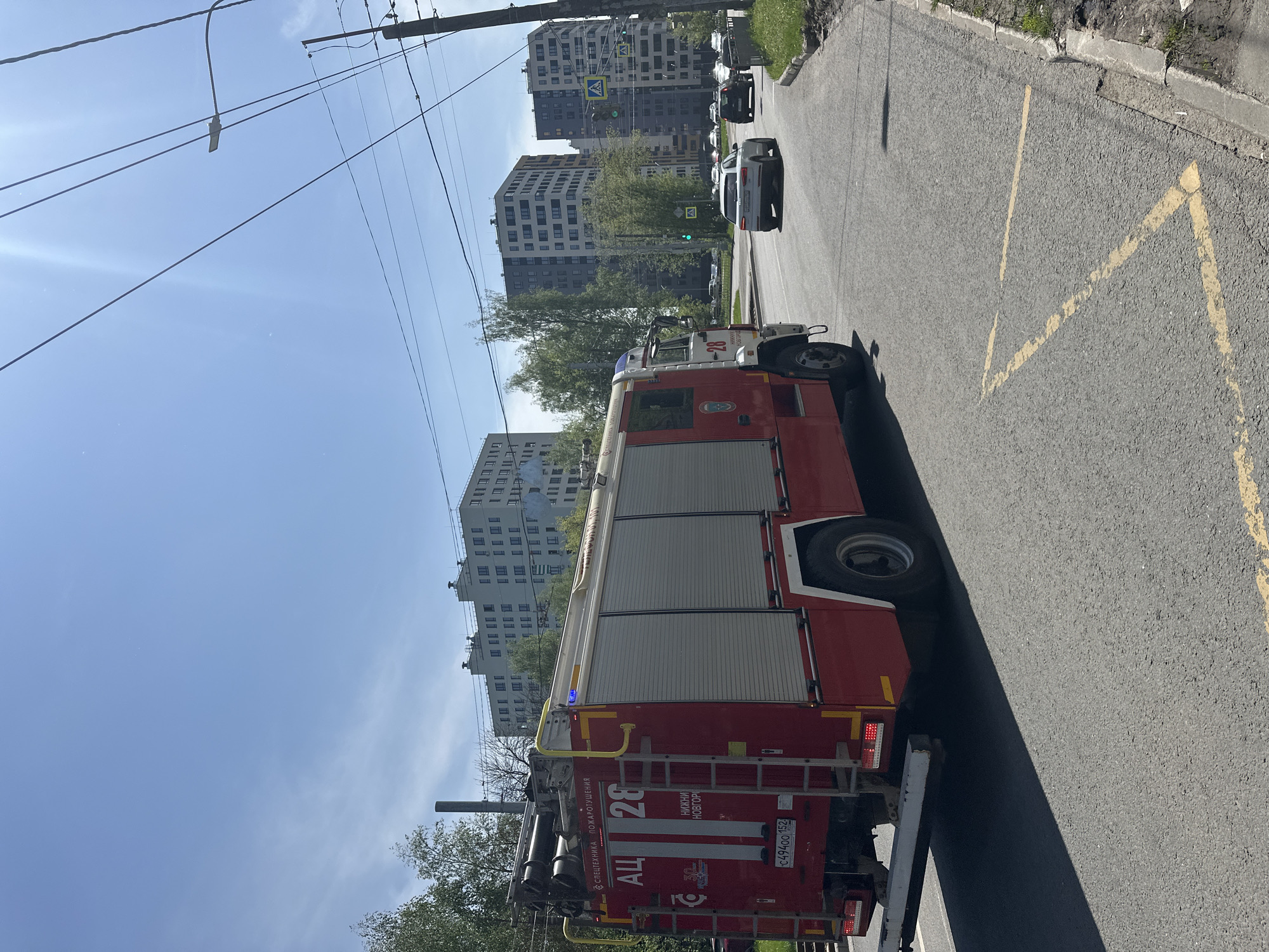 The image shows a fire engine parked on a street in Nizhny Novgorod, Russia. The fire engine is red and has "АЦ" (which is a type of fire engine in Russian) and "Нижний Новгород" (Nizhny Novgorod) written on its side. The number "26" is visible on the back. The registration number "C494OO 152" is visible on the license plate.
Several apartment buildings are in the background. The weather appears to be clear. Other vehicles are visible further down the street. Yellow markings are on the road. Overhead power lines are visible.