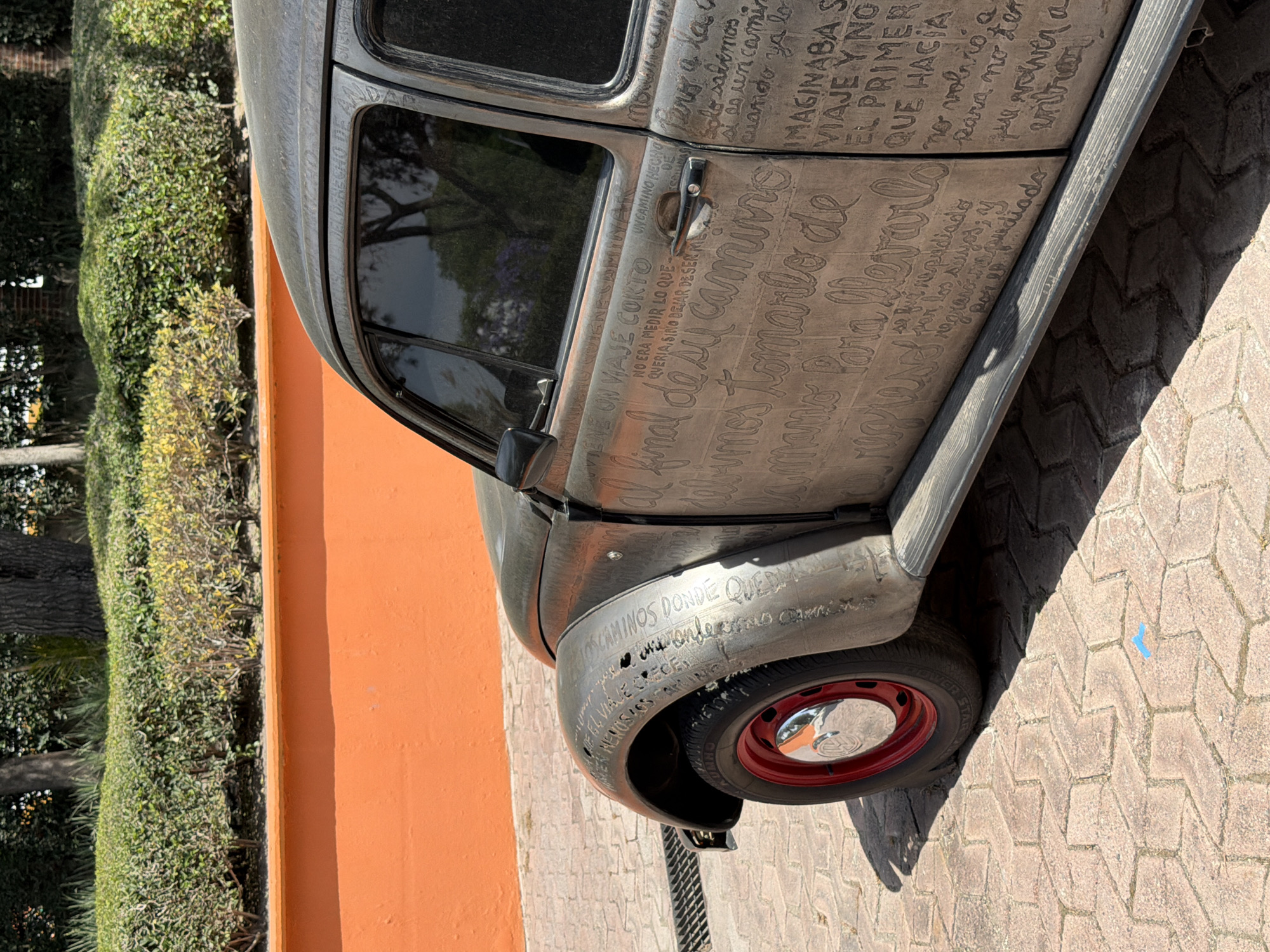 The image showcases a modified classic Volkswagen Beetle, positioned on a brick-paved area next to an orange wall and a lush green hedge. The car's body is adorned with etched text in Spanish, covering most of its surface. The Beetle has a red-rimmed wheel with a chrome hubcap. The setting is in San Andrés Cholula, Mexico, although specific landmarks are not visible to confirm the location within the city. The scene appears to be a display or exhibition of the unique Beetle.