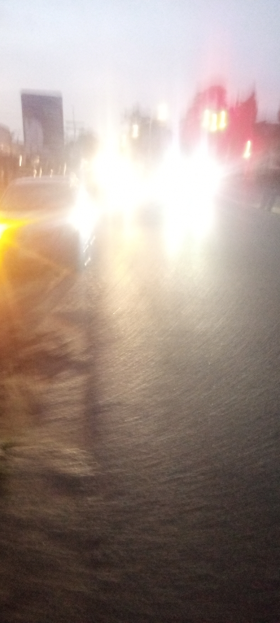 The image shows a street scene at dusk or night in what appears to be Kabala, Nigeria. Bright headlights of oncoming vehicles illuminate the wet road, obscuring details of the cars themselves. To the left, a vehicle with yellow lights is visible. In the background, indistinct buildings and a billboard can be discerned. It is either raining or the road is wet from recent rain, reflecting the lights and obscuring the texture of the road surface.