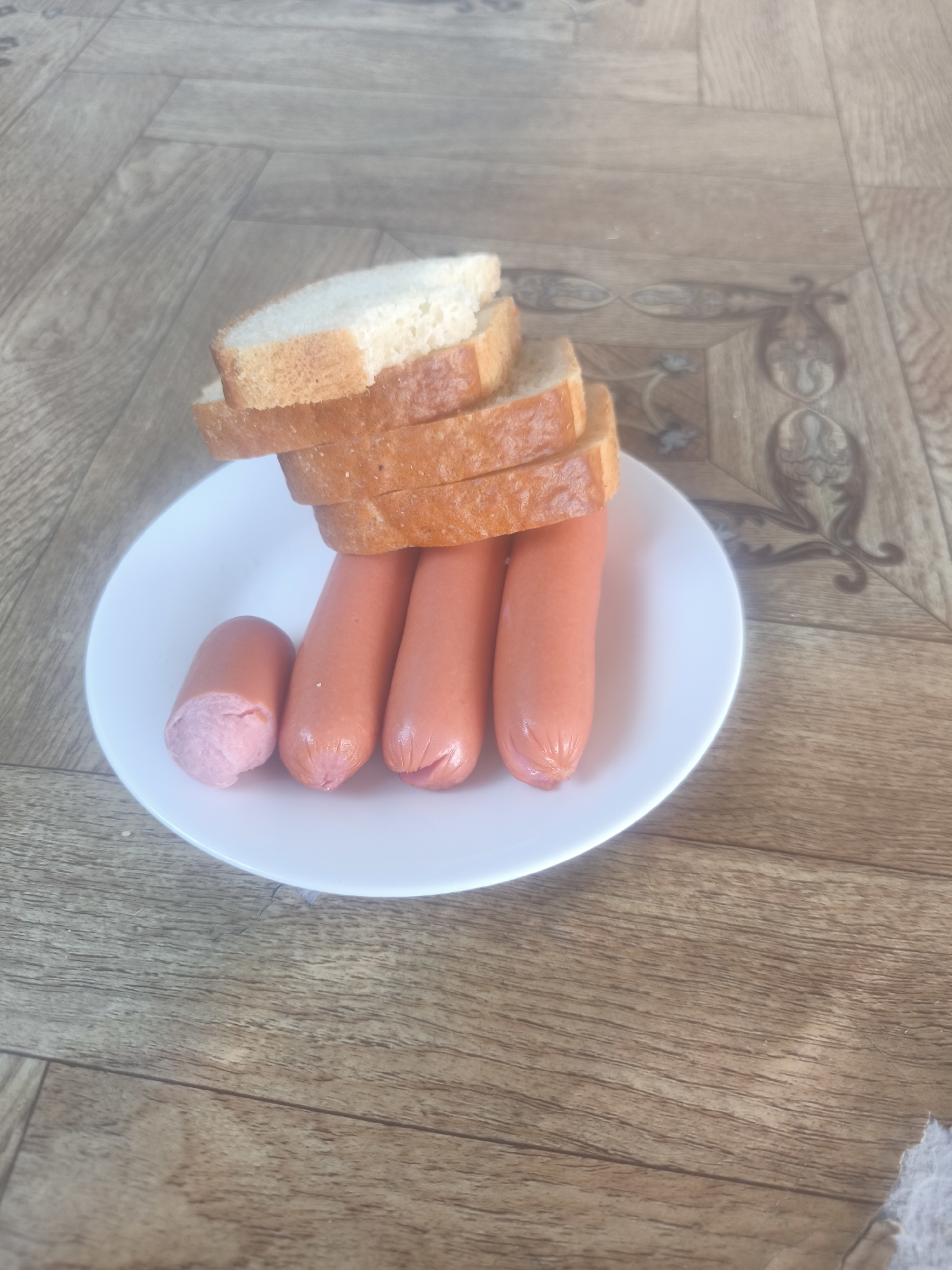 This image depicts a white, round plate resting on a patterned floor surface, containing four frankfurters and four slices of white bread. Three of the frankfurters are whole, appearing pinkish-orange with smooth casings. The leftmost frankfurter is bisected, revealing a lighter pink, textured interior. Stacked vertically atop the frankfurters are four slices of white bread with golden-brown crusts; the topmost slice exhibits a small tear or bite mark on its right edge. The background surface is a light-brown, wood-grain patterned linoleum or laminate flooring, featuring darker brown decorative inlays with ornamental designs, particularly noticeable in the upper and lower right quadrants of the frame. The scene is evenly lit. No individuals are present. Location: Unknown, Unknown.
