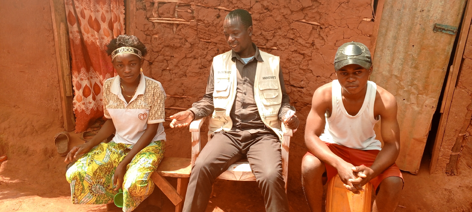 A wide shot depicts three individuals – a young woman and two men – seated outdoors against a textured, reddish-brown earthen wall in Ngalula, Democratic Republic of the Congo. The ground is also bare earth.
On the left, a young woman with dark, curly hair wears a white headband featuring the text "GUCCI" in gold. She is dressed in a light-colored polo shirt with a patterned print and a long, vibrant yellow and green patterned skirt. A green plastic cup is visible near her feet, and brown sandals are behind her. Behind her, a fabric curtain with a brown leaf pattern hangs, likely covering an entrance or window.
In the center, an adult man sits on a white wooden chair. He wears a dark long-sleeved shirt and a light-beige multi-pocket vest, which clearly displays the text "ANOVEV-EF3" on two of its pockets. He appears to be speaking or explaining something, gesturing with his right hand.
On the right, a young man sits on an orange plastic bucket or stool. He wears a dark baseball cap with a silver-colored logo, a plain white sleeveless undershirt, and red shorts. He looks forward with his hands clasped together. Behind him, a corrugated metal sheet, possibly a door, is partially visible.
The scene is brightly illuminated by natural sunlight, indicating it is daytime. The earthen architecture, simple seating, and attire suggest a rural setting. The individuals exhibit neutral to slightly serious expressions, suggesting a conversation or interview is underway.