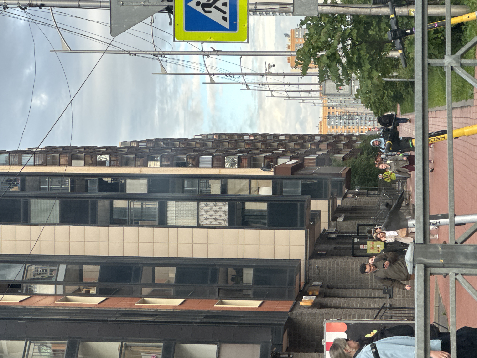 This is a vertical, eye-level, medium shot of a street scene in what appears to be Russia, given the Cyrillic text on a sign. The sky is overcast, suggesting it's likely daytime with cloudy weather.
The primary subject is a modern, multi-story building with a beige and dark-paneled facade. Many windows are visible, some reflecting the sky and others showing glimpses of interior spaces. A patterned curtain is visible in one window. The building's architecture is characterized by repetitive, rectangular window units.
In the foreground, a dark metal fence runs vertically along the right side of the frame, partially obscuring the view of a brick wall and a sidewalk paved with reddish-brown bricks. A portion of a yellow scooter is visible, with the word "JOSH" printed on it.
Several people are present on the sidewalk, some walking and others standing. One person is wearing a dark hooded jacket and appears to be looking at their phone. Another person in the foreground is wearing a light blue jacket and looking up.
Above the building, a network of overhead wires and poles stretches across the frame, typical of urban infrastructure. A street sign with a blue background and a white pedestrian symbol is visible near the top left.
The overall impression is of a typical urban environment, likely in the late afternoon or evening, given the diffused light. The scene conveys a sense of everyday life in a city.