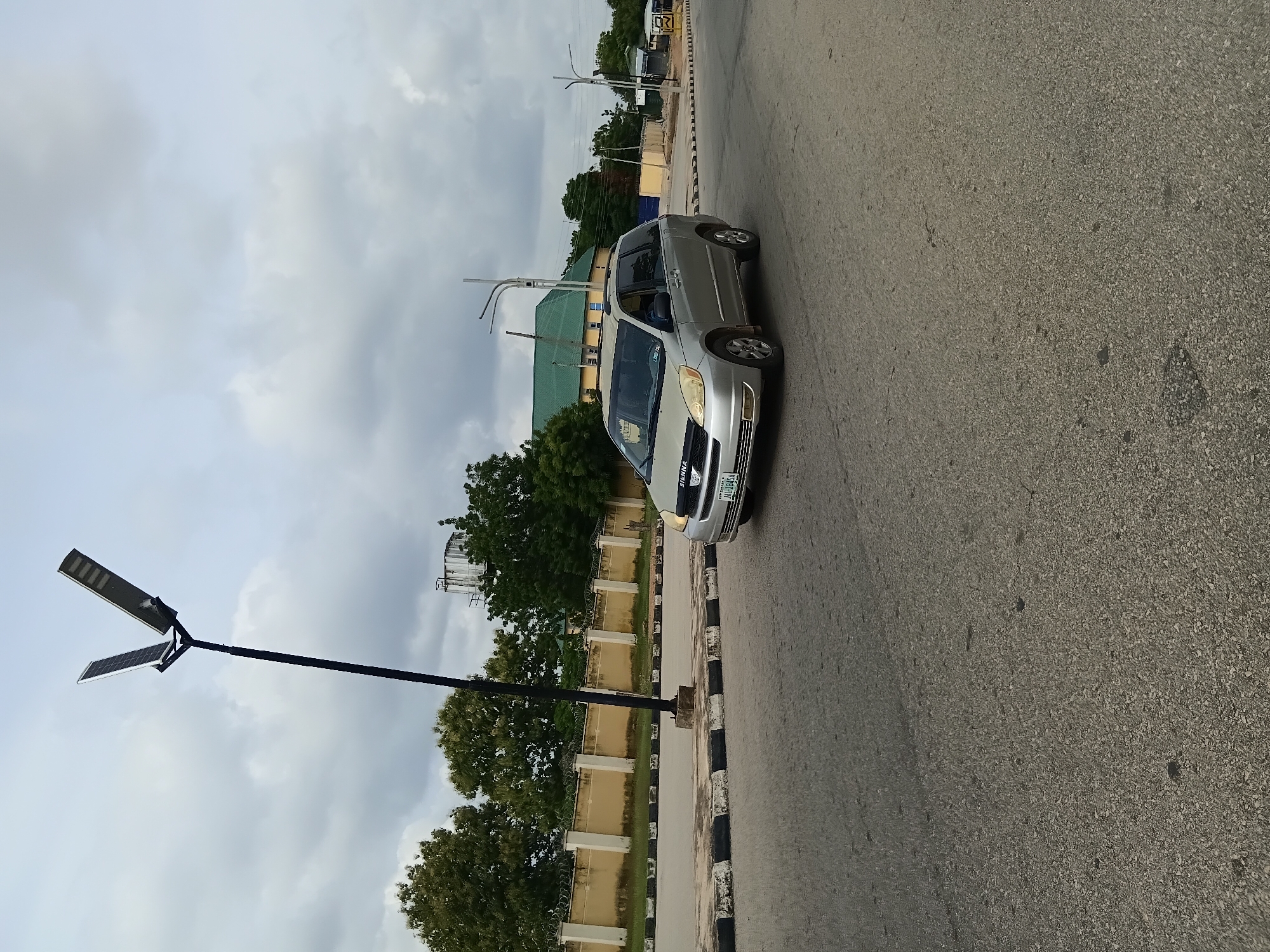 The image shows a silver minivan, identified as a Toyota Sienna, driving on a paved road in Jalingo, Nigeria. The minivan has a license plate that reads "JAC 1895A". The road has a black and white striped curb. In the background, there is a yellow wall with barbed wire on top, behind which are several trees. A solar-powered street light is prominently positioned on the left side of the image, with its panels angled upwards. Further in the background, more trees and buildings are visible, including a structure with a green roof and what appears to be a water tower. The sky is cloudy, indicating overcast weather. The scene appears to be an urban environment in Jalingo, Nigeria, with a focus on the road and traffic.