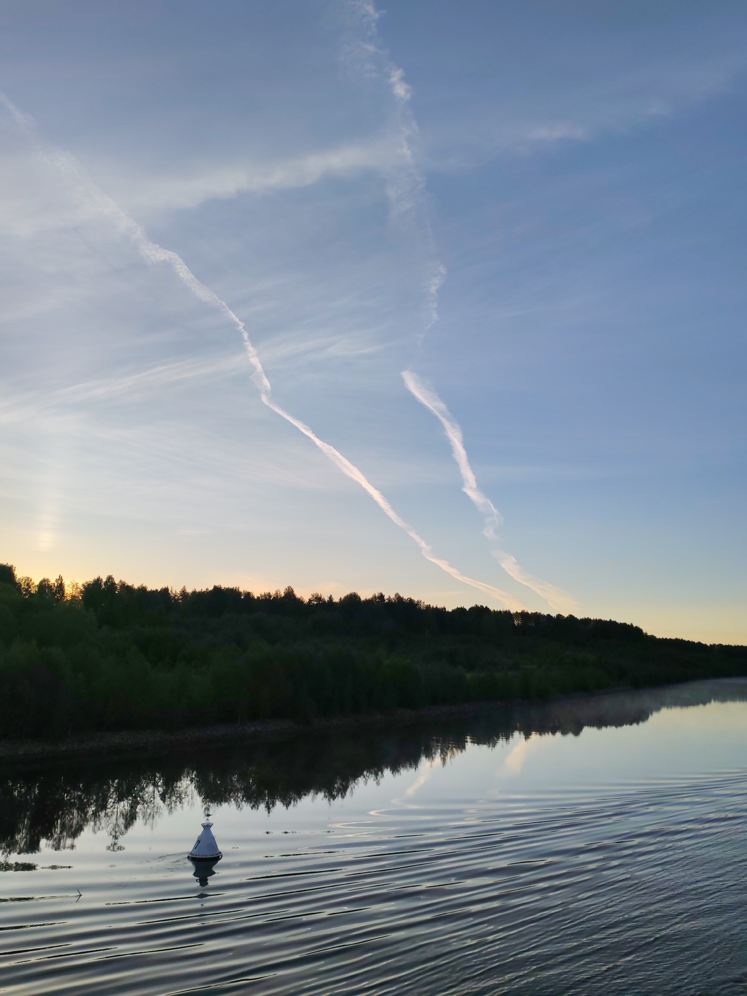 The image presents a tranquil landscape scene at what appears to be either sunrise or sunset over a body of water in Volokov Most, Russia. The sky dominates the upper half, displaying a gradient from a soft, warm yellow-orange hue near the horizon on the left, transitioning to a pale blue overhead towards the right. Several prominent, white aircraft contrails cut across the sky; one particularly long, wavy trail extends from the upper right, intersecting with another fainter trail closer to the upper left.
A dense, dark band of trees or forest lines the distant bank, casting a clear reflection onto the remarkably calm water below. The water occupies the lower half of the image, serving as a reflective surface for both the trees and the sky. In the lower left quadrant of the water, a white, conical buoy with a small dark top floats, creating subtle ripples around it and casting a dark reflection. More distinct, parallel wave patterns are visible across the water in the foreground, stretching from the right side towards the center.
The scene conveys a sense of peacefulness and natural beauty, with the atmospheric contrails adding a modern touch to the serene vista. There are no people visible, and no discernible text.