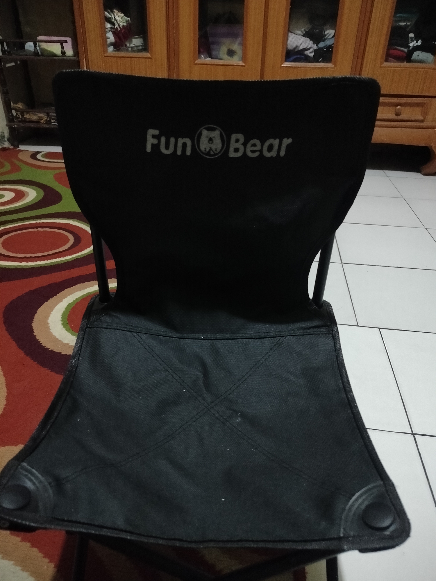 A black folding chair with a "Fun Bear" logo is centered in the frame, positioned on a colorful, circular-patterned rug. The chair is made of fabric and metal, with a wide seat and a slightly curved backrest. The logo, featuring a bear's face within a circle, is visible on the upper portion of the chair's backrest. The background includes a wooden cabinet with glass doors, revealing stored items, and a tiled floor. The scene appears to be indoors, likely in a residential setting in Kumbangindah, Indonesia.