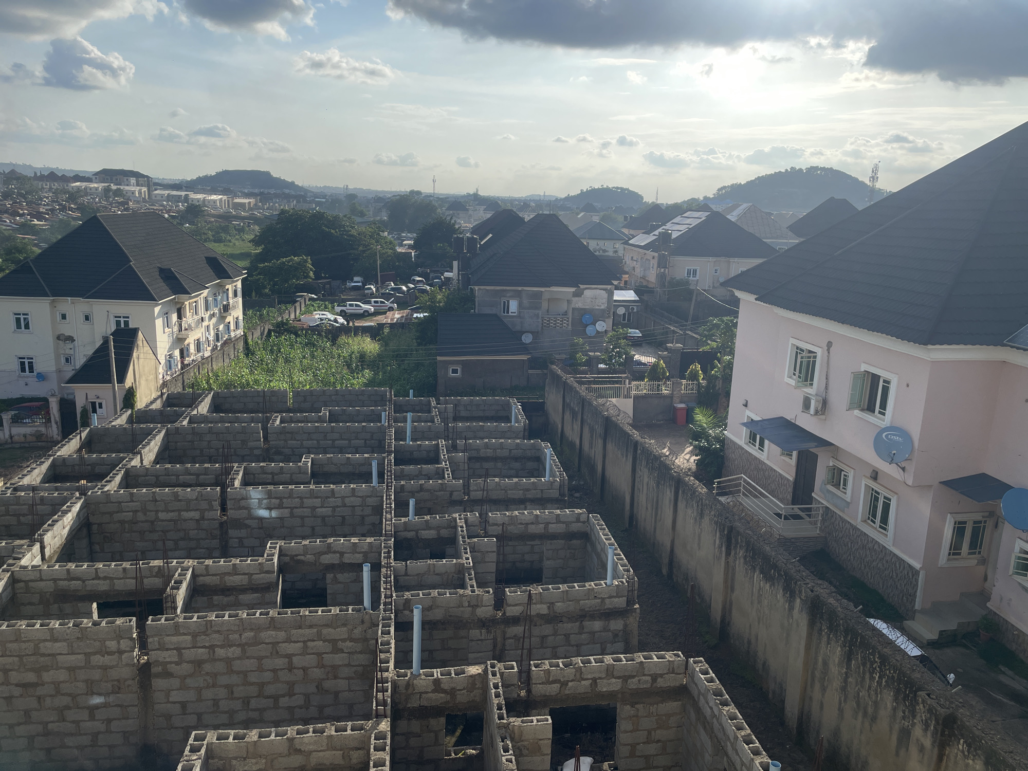 The image shows a developing residential area in Abuja, Nigeria, as viewed from an elevated position. In the foreground, the foundations of multiple houses are under construction, characterized by concrete block walls forming various room outlines. Several PVC pipes protrude vertically from the unfinished structures. A tall concrete wall, topped with barbed wire, separates the construction site from a neighboring property.
Beyond the construction, the scene opens up to a cityscape with a variety of houses, many featuring dark-tiled roofs. Some of these houses appear completed and inhabited, while others are still in various stages of construction. In the middle ground, a parking lot is visible with several cars. The background reveals a hilly landscape, characteristic of Abuja's topography, with some structures and vegetation on the slopes. The sky is partly cloudy, with sunlight filtering through, suggesting it is daytime. The overall impression is of urban expansion and development within the city.