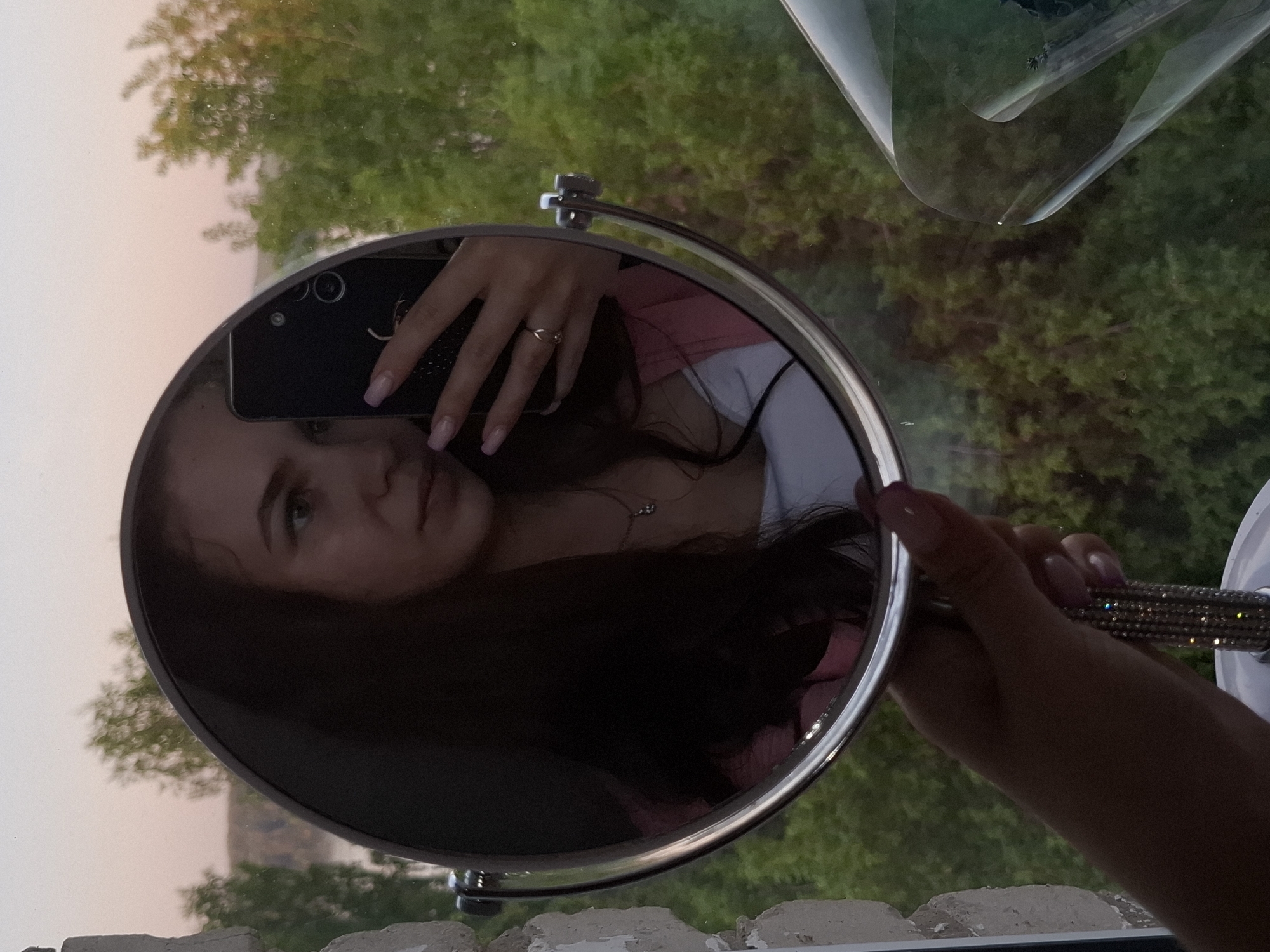 A woman is holding a round mirror, taking a selfie with her phone in the mirror’s reflection. The reflection shows her face, part of her upper body wearing a white top and a pink garment, her hand holding the phone, and a ring on one of her fingers. The mirror has a silver frame and a handle decorated with rhinestones. Outside the window, there is green foliage and a glimpse of the sky, indicating an outdoor setting. The location is Chaikovsky, Russia.