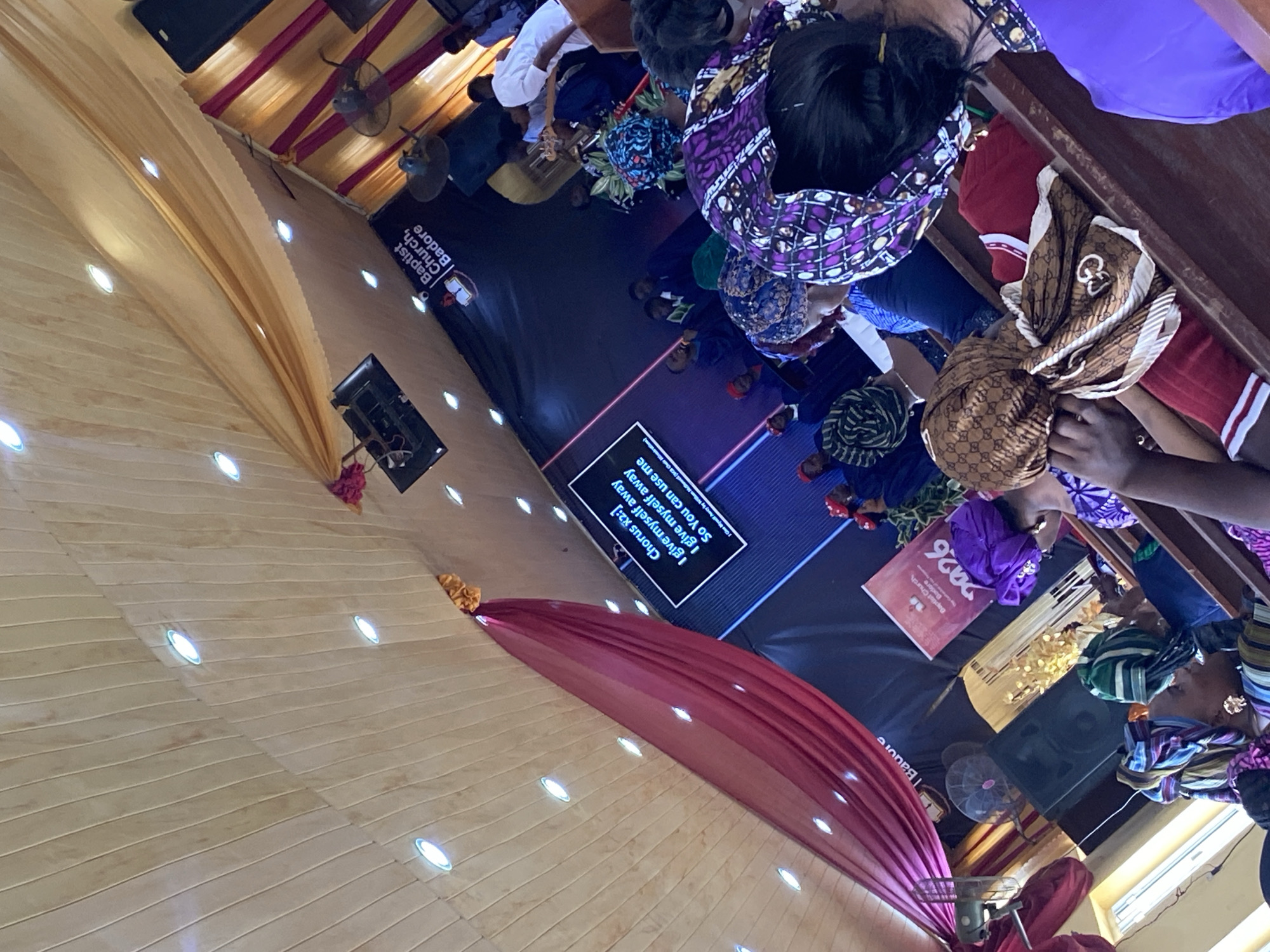 The image depicts the interior of the Baptist Church, Badore, likely located in Lagos, Nigeria, based on the signage visible. A mixed group of people, predominantly women, are attending a service. Many are wearing colorful African headwraps. A screen displays the lyrics of a song: "Chorus x2: I give myself away, I give myself away, So You can use me." The decor includes golden and pink draping along the ceiling and walls.